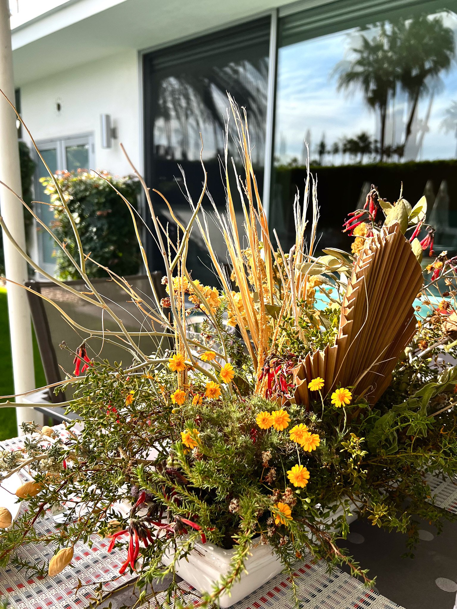A vibrant floral arrangement featuring yellow, red, and green plants in a white rectangular container, set on a patterned table outdoors with a modern house and a swimming pool in the background.