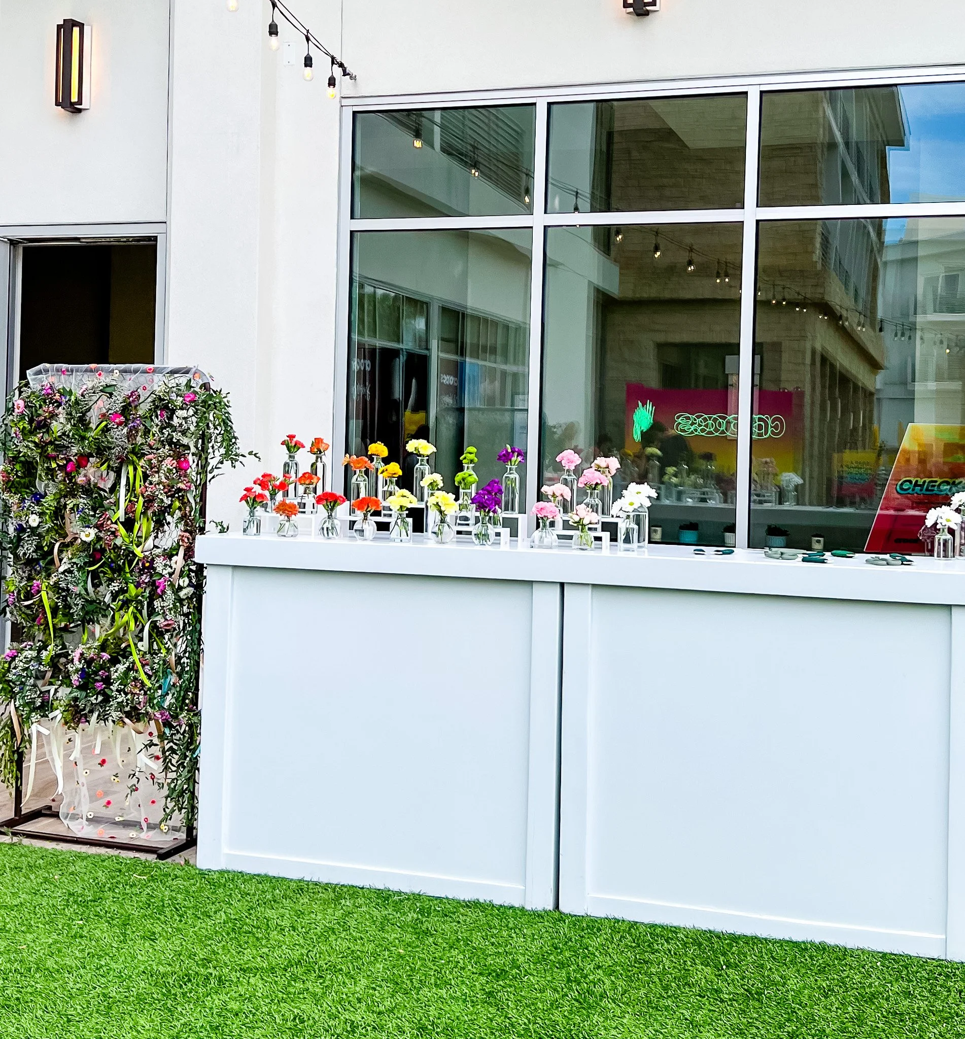 Bud vases filled with flower stems arranged by the colors of the rainbow. The bud vases are on a white bar top. There are scissors and wire on the bar top too. To the left of the bar, there is a display of hanging flower crowns.