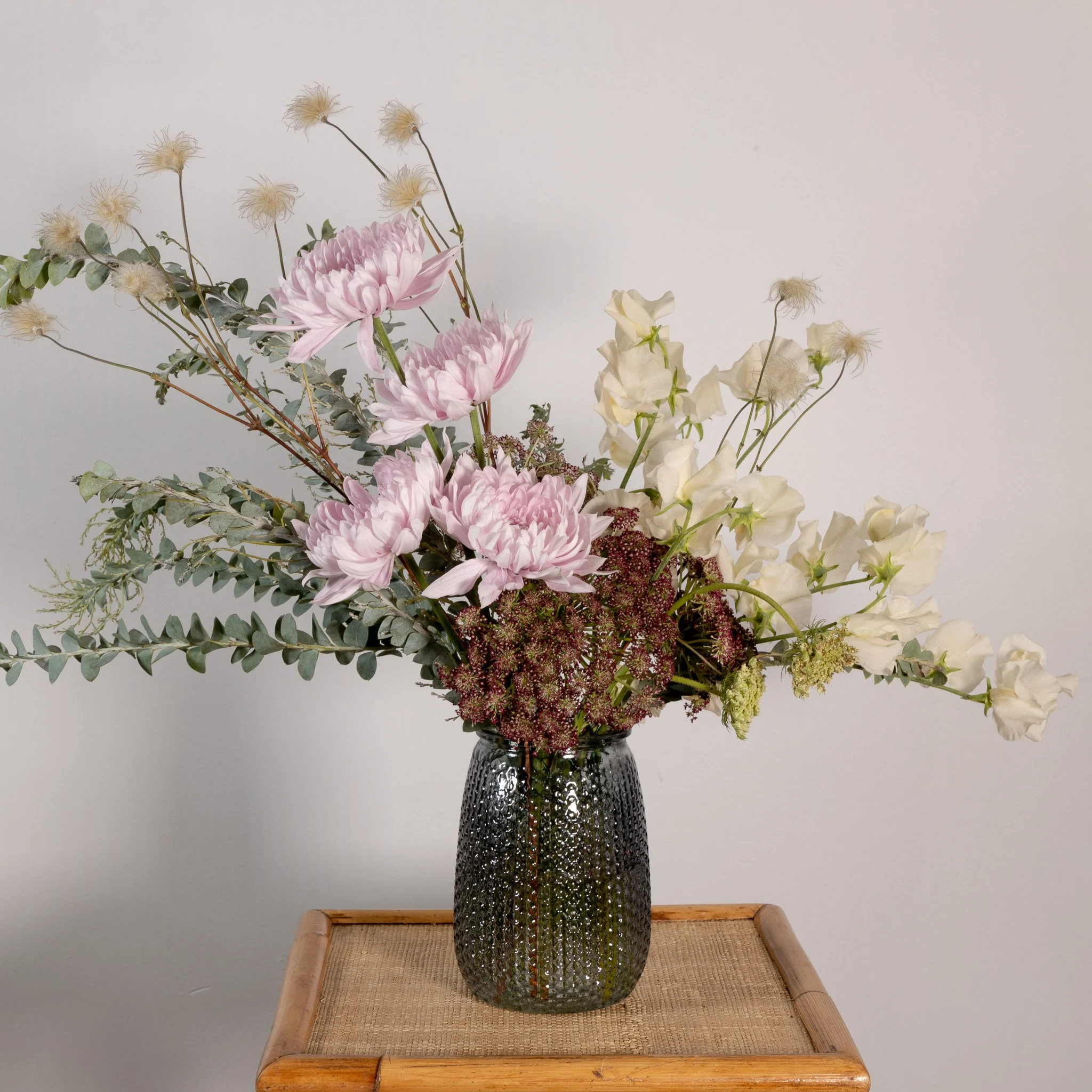 A flower arrangement with pink chrysanthemums, white sweet peas, and beige fluffy flowers in a black textured glass vase on a wooden table.