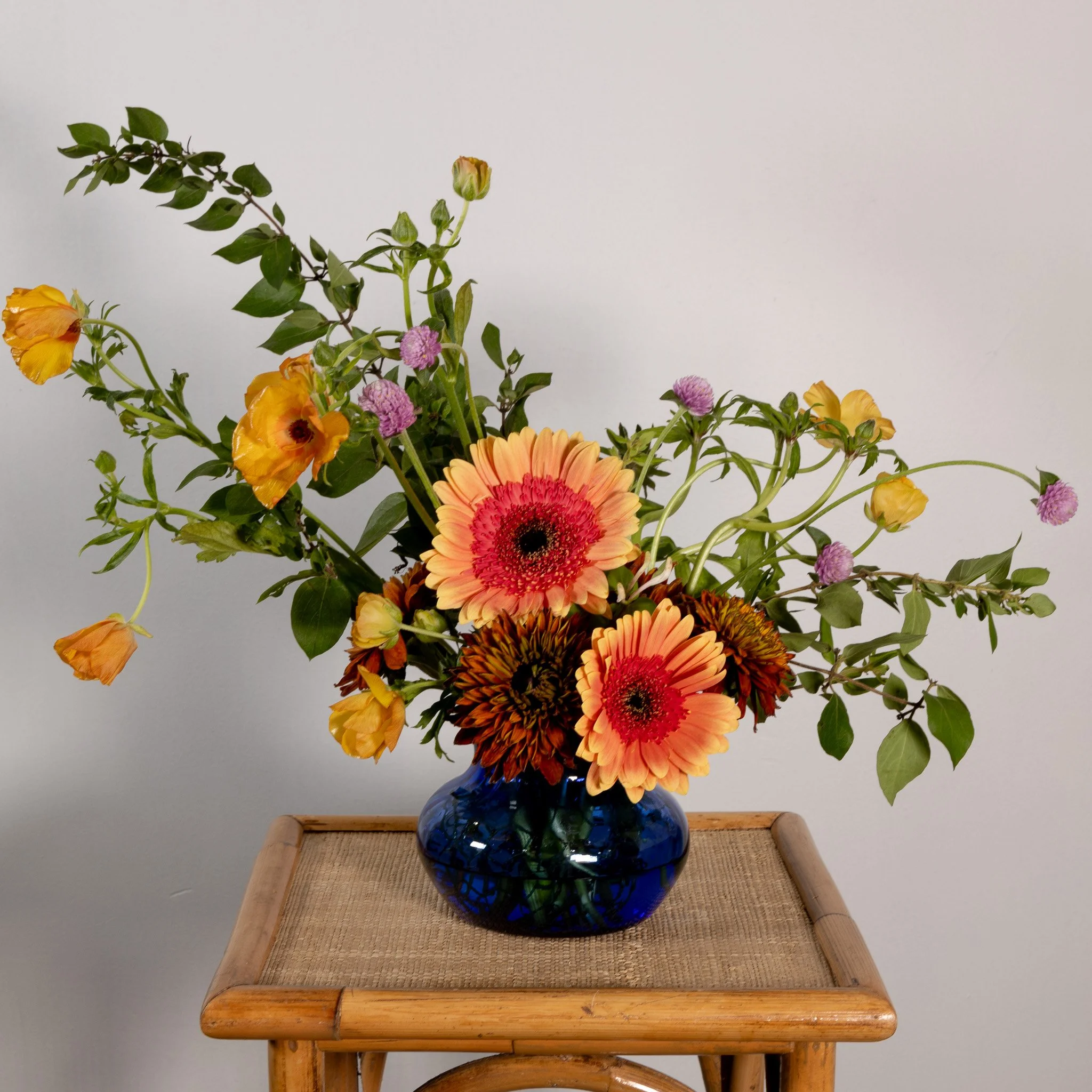 A colorful flower arrangement in a blue glass vase on a wooden table against a plain white wall.