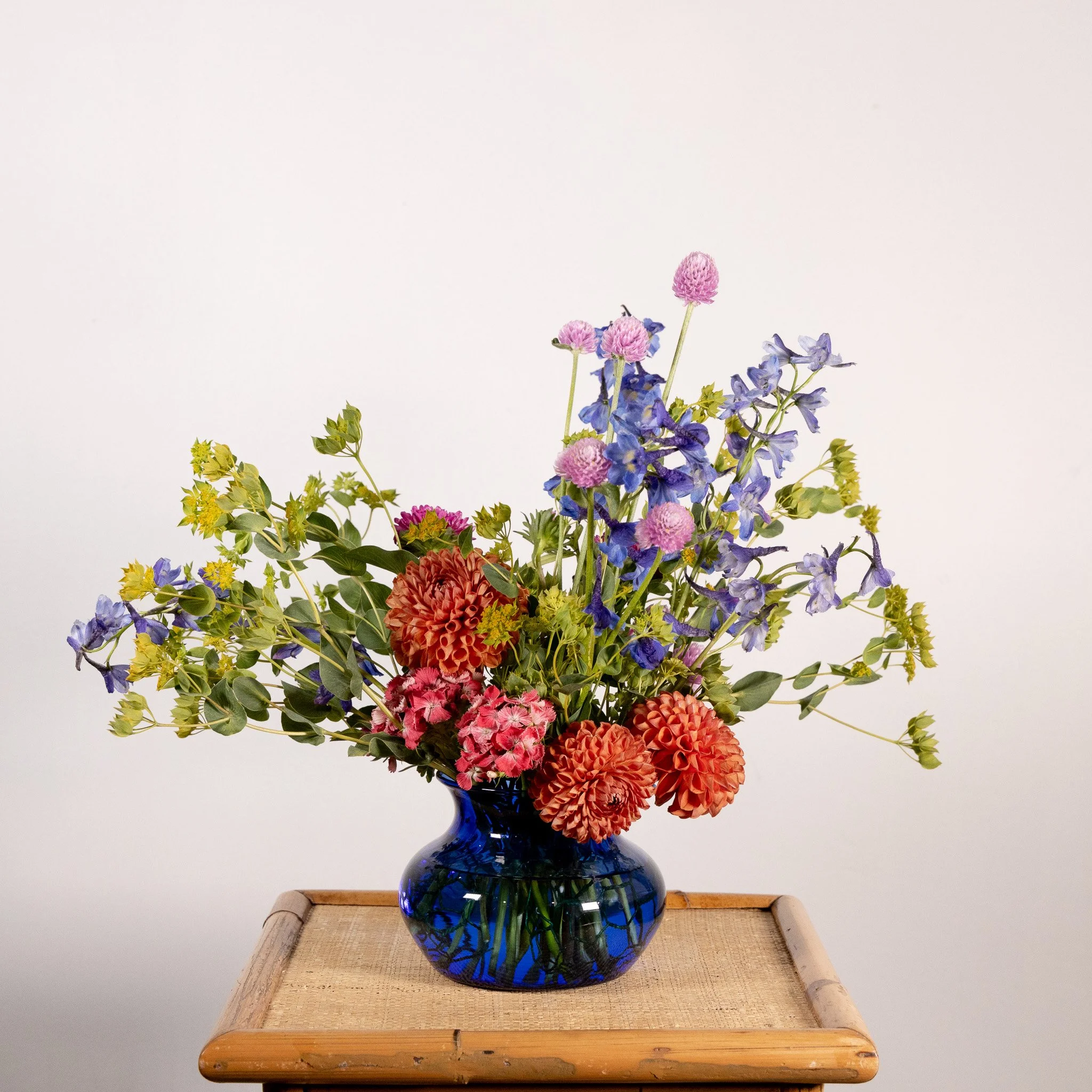 A colorful bouquet of flowers, including dahlias, delphiniums, and pink pom-poms, arranged in a deep blue glass vase on a wooden table against a plain white background.