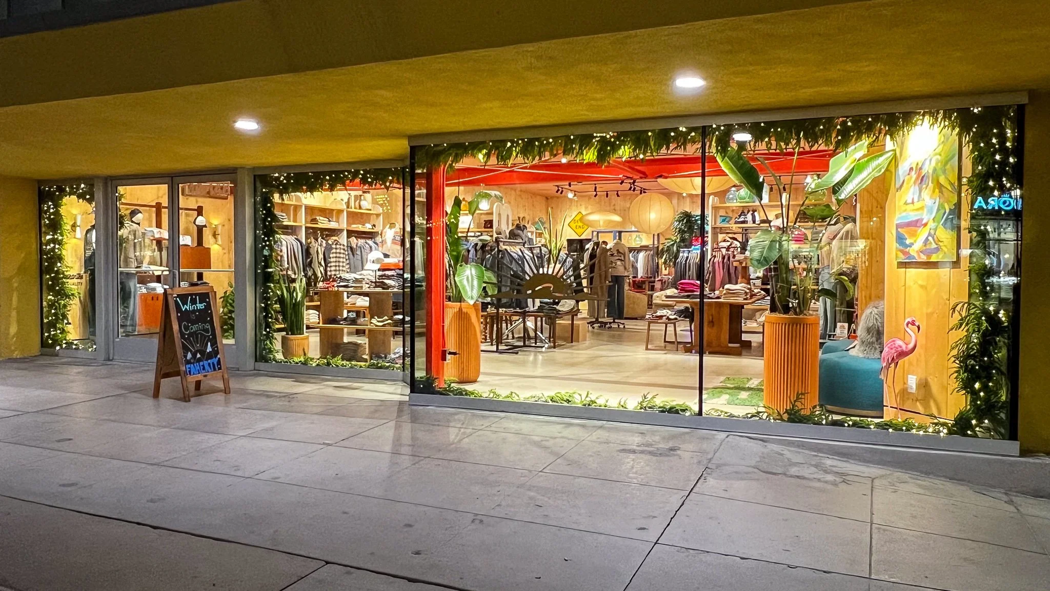 Clothing store window at night decorated with greenery and fairy lights, with a sign outside advertising winter clearance, displaying various clothing and accessories inside.