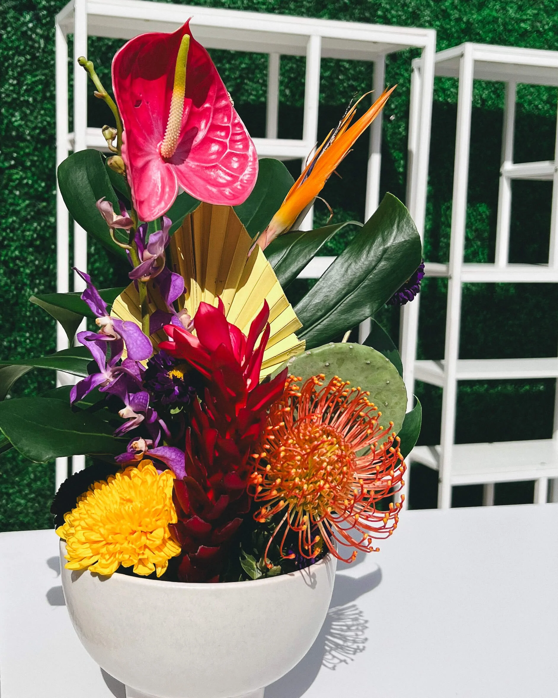 A bright colorful arrangement sits on top of a bar. It features tropical and desert-related plants, like anthuriums, orchids, monstera leaf, cactus paddle, pincushion protea, ginger flower, and a mum.