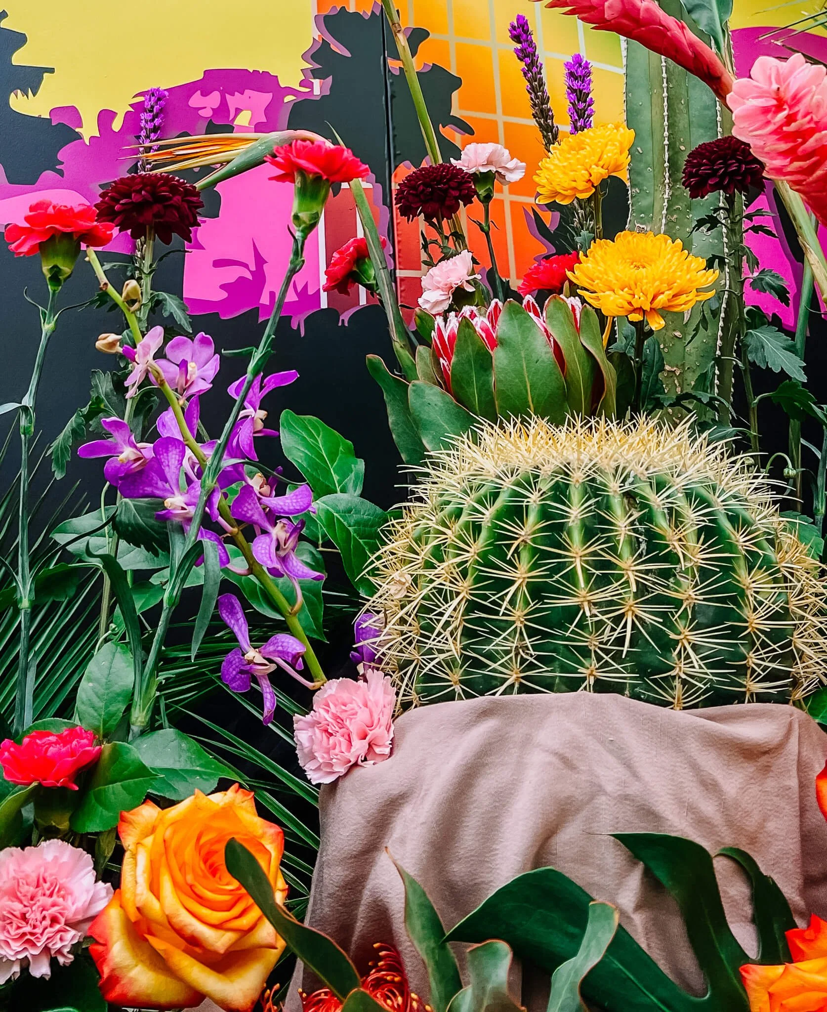 A golden barrel cactus's pot is covered by sand-color fabric while bright flowers are surrounding the cactus.