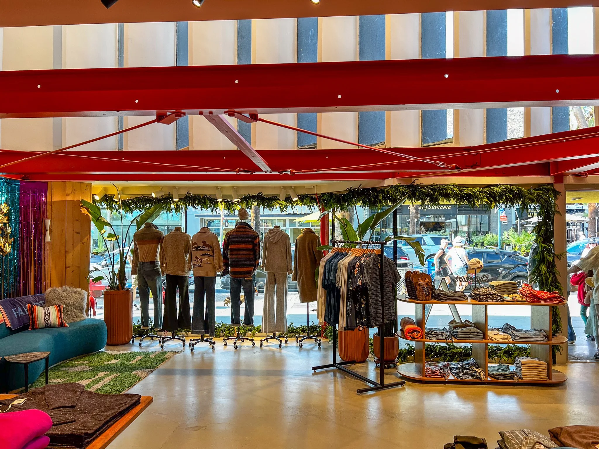 Inside a clothing store with mannequins dressed in sweaters and pants near the front window, racks of clothes, and decorative plants, with large windows showing pedestrians walking outside.