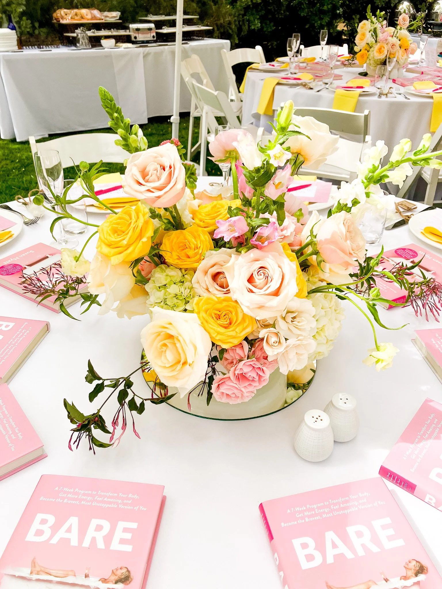 Elegant table setting at a daytime event with a centerpiece of pink, yellow, and peach roses and flowers, pink event booklets, and white salt and pepper shakers, outdoors with other decorated tables in the background.