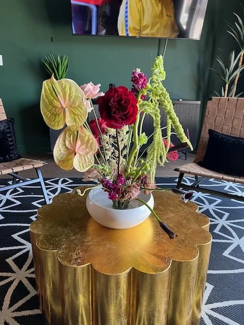 A white bowl with various colorful flowers, including sizes of pink, red, and green, sits on a gold, scalloped table in a room with black and white patterned carpet, black pillows, and potted green plants.