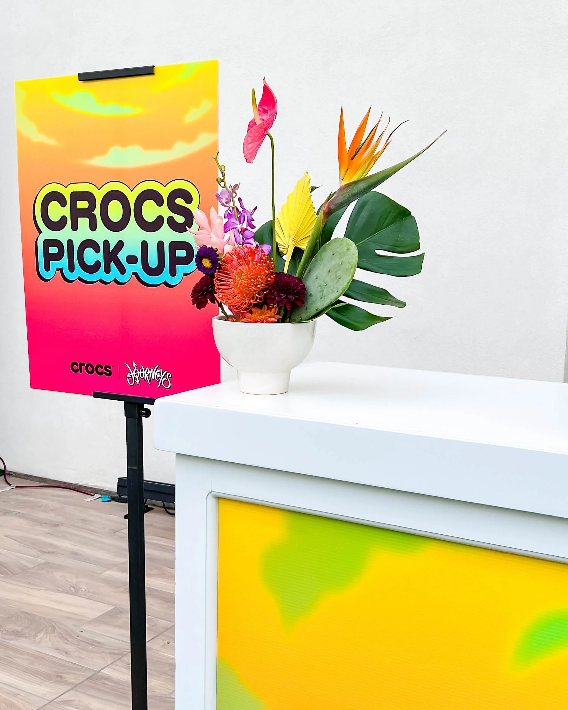 A bright colorful arrangement sits on top of a bar. Event brand signs for Crocs and Journeys is next to it.