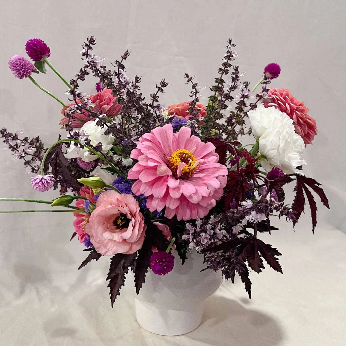 Medium sized flower arrangement for weekly subscriptions with purple and pink flowers and foliage in a modern white vase.
