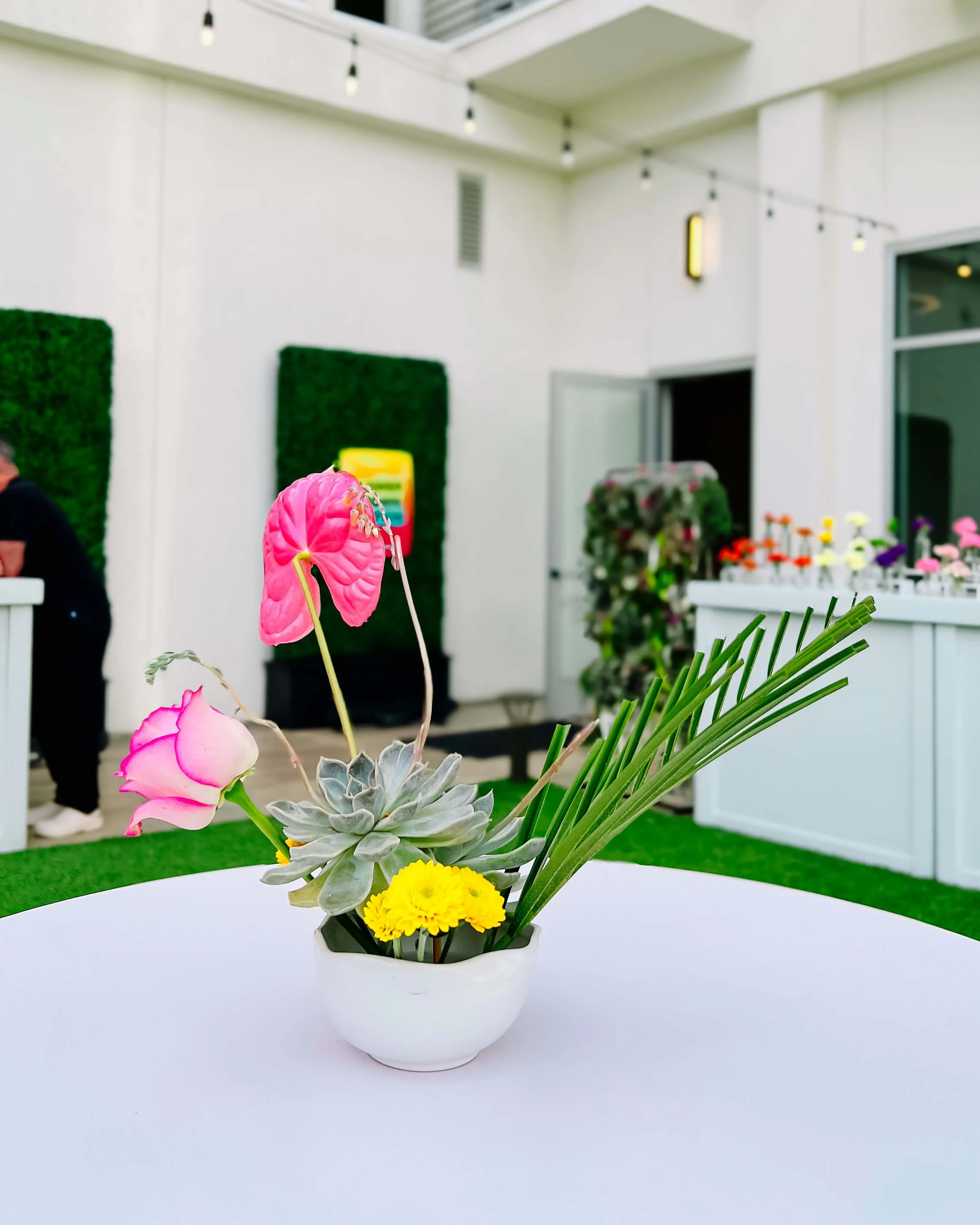 Small cocktail arrangement in a white bowl with a pink anthurium, rose, palm frond, succulent, and button mums. It's on a white, round cocktail table and a flower crown station is in the background.