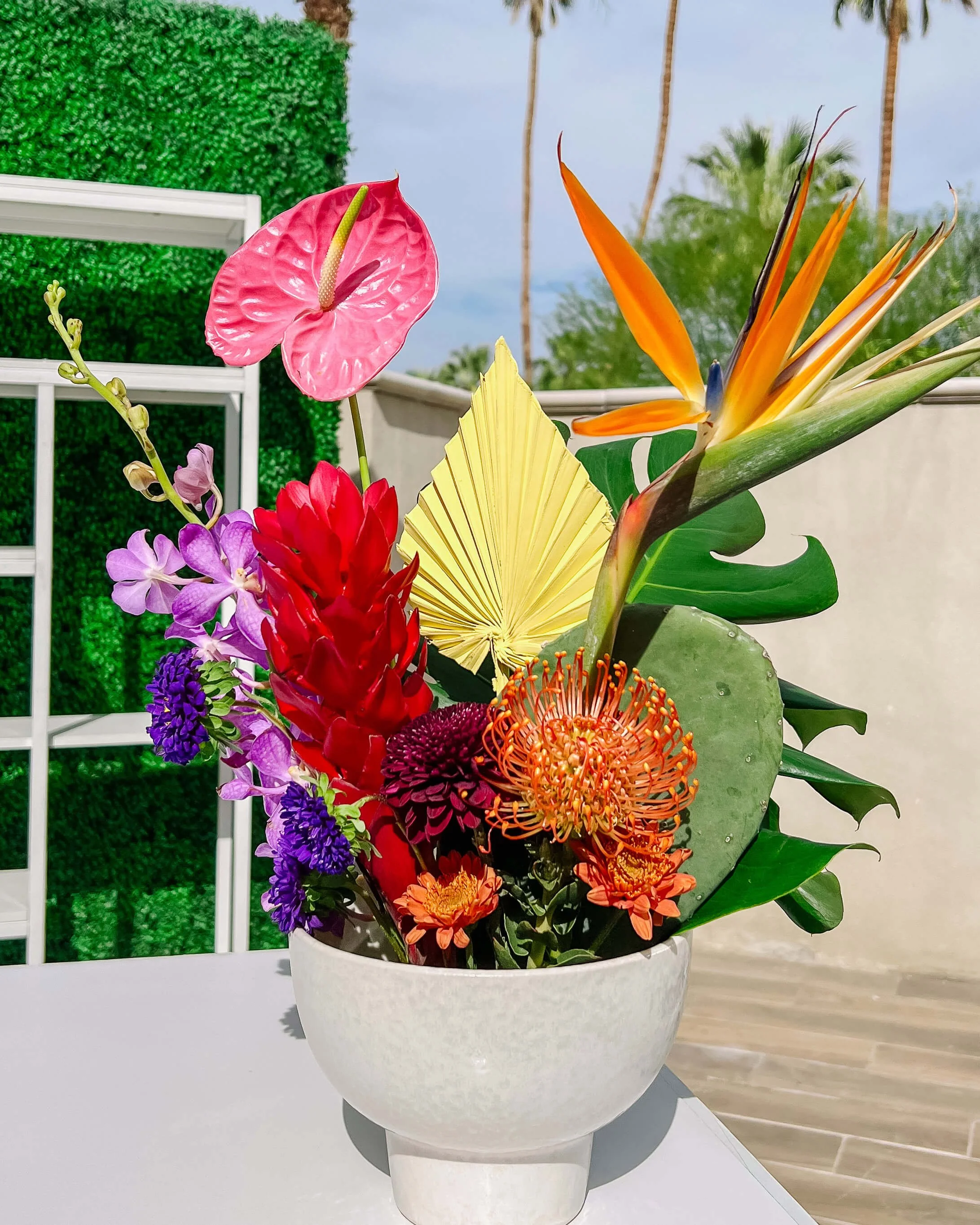 A bright colorful arrangement sits on top of a bar. It features tropical and desert-related plants, like anthuriums, orchids, monstera leaf, cactus paddle, pincushion protea, ginger flower, asters, bird of paradise, and a painted palm spear.