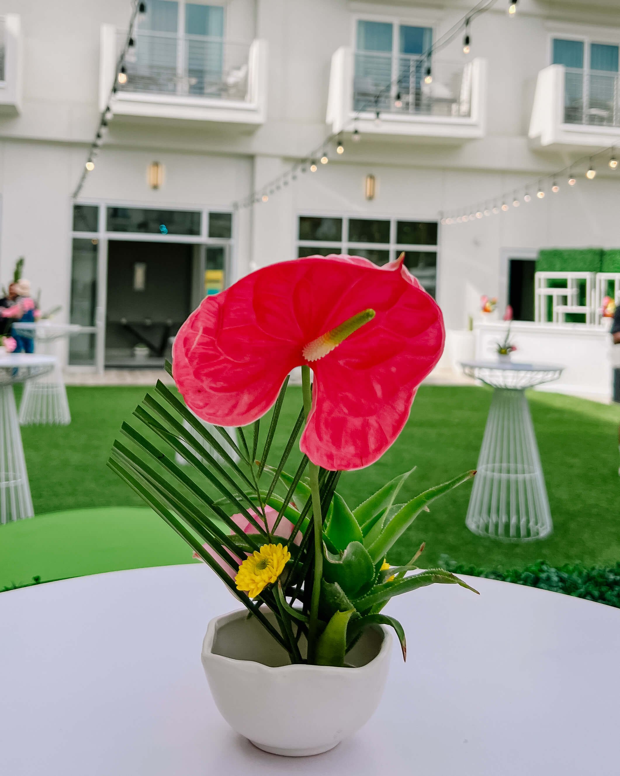 Small cocktail arrangement in a white bowl with a pink anthurium, rose, palm frond, aloe, and button mums. It's on a white, round cocktail table and other cocktail tables are in the background on a green lawn.