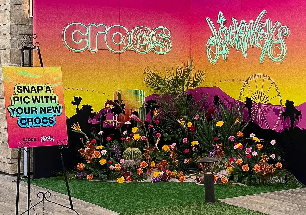 A large photo backdrop floral installation that shows the various bright colored flowers, succulents, a palm tree, palm fronds, and kinetic sand that were used in the design. A sign on an easel next to the install reads Snap a pic with your new Crocs