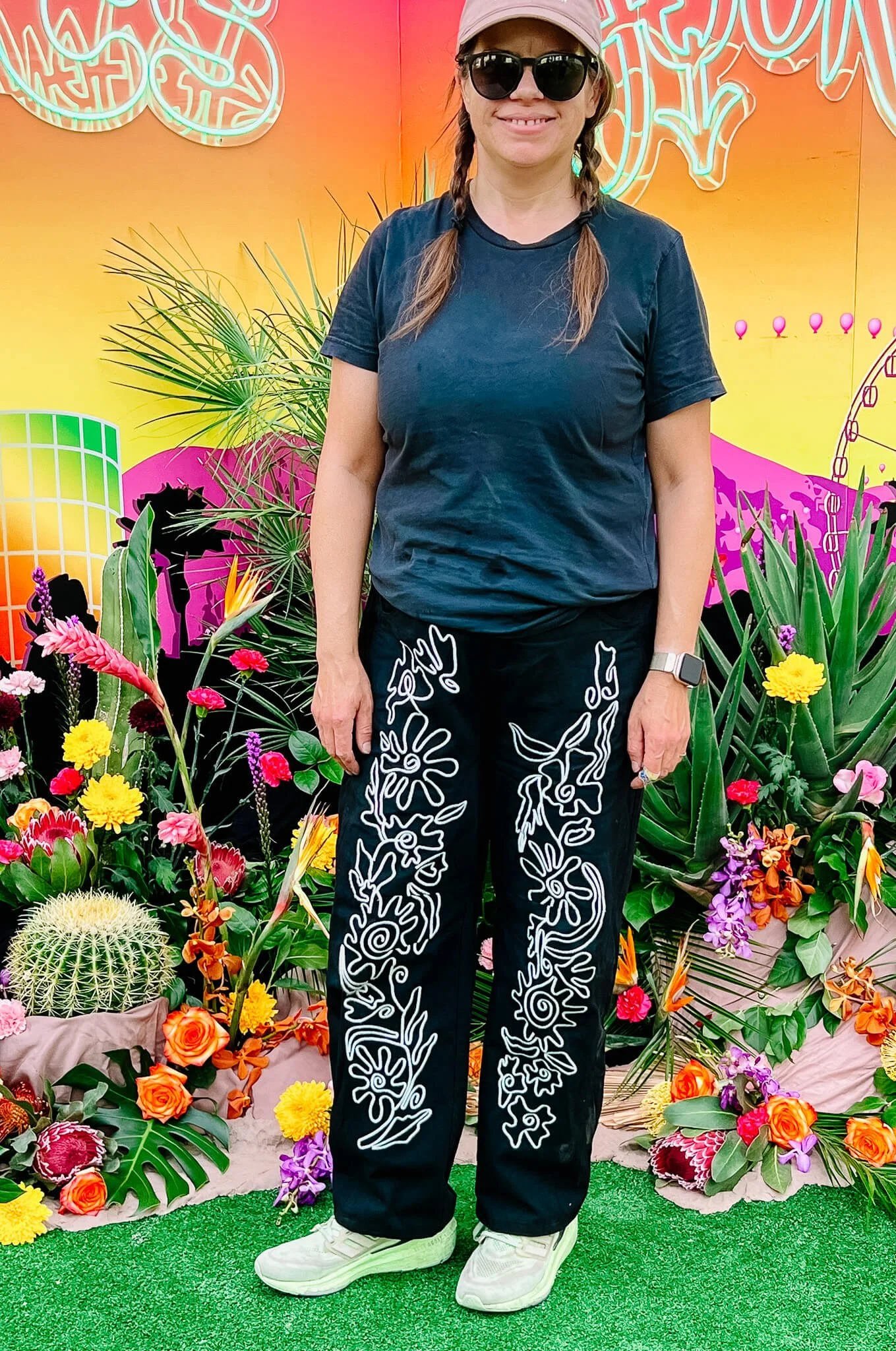 A woman in a tshirt, jeans with embroidered abstract flowers, sunglasses, and a hat stands in front of the botanical installation.