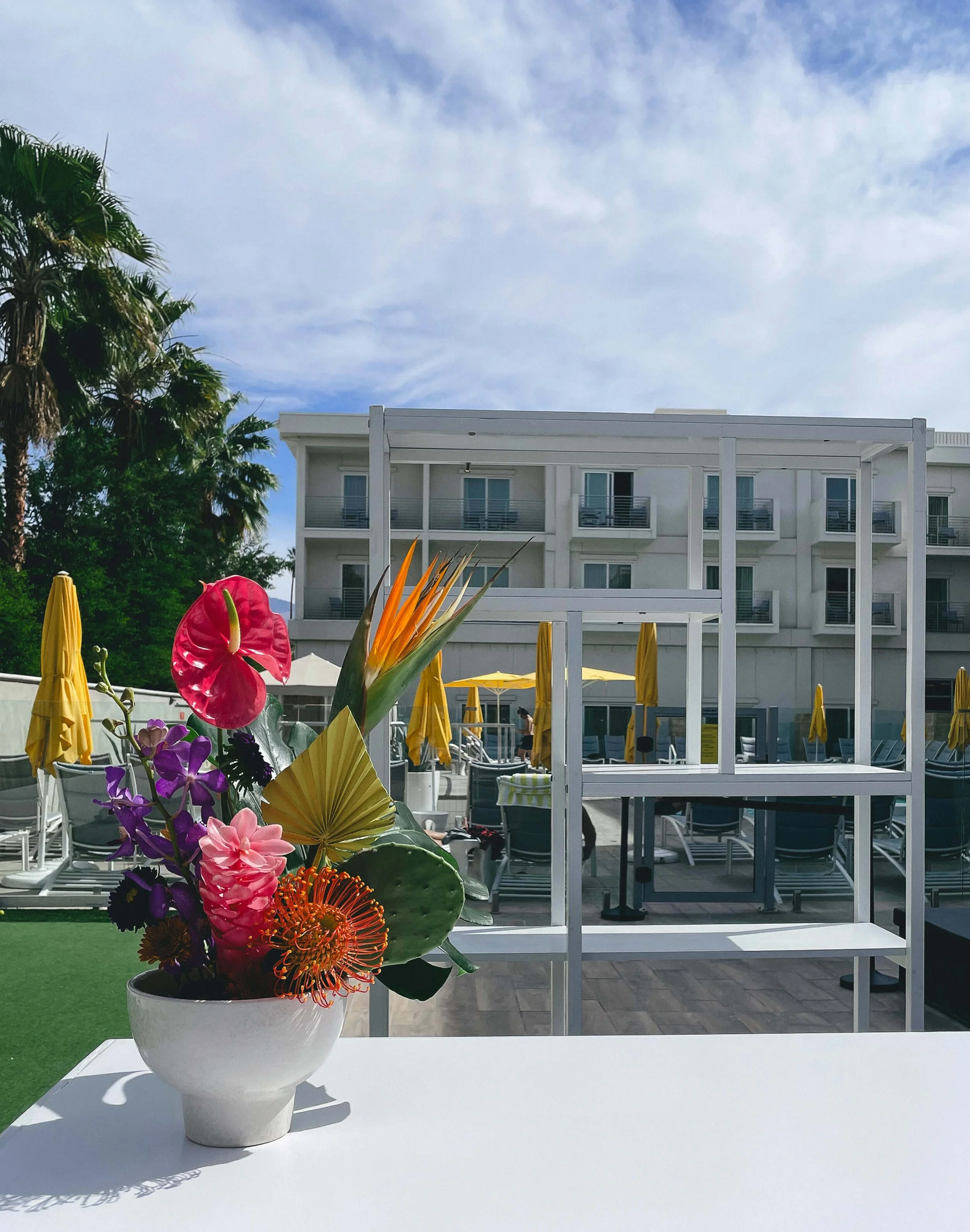 A bright colorful arrangement sits on top of a white bar top. Patio chairs for a pool and umbrellas are in the background.