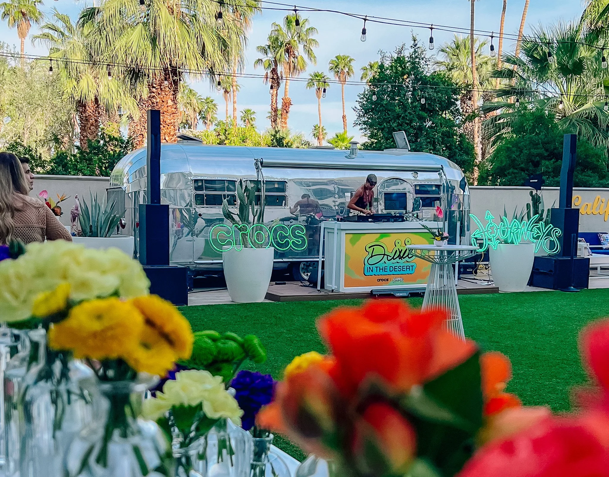 A DJ standing at a DJ booth in front of an airstream. Neon signs of the Crocs and Journeys logos are on either side of the booth resting on top of large potted cacti. In the foreground are out of focus bud vases of flowers for a flower crown station.