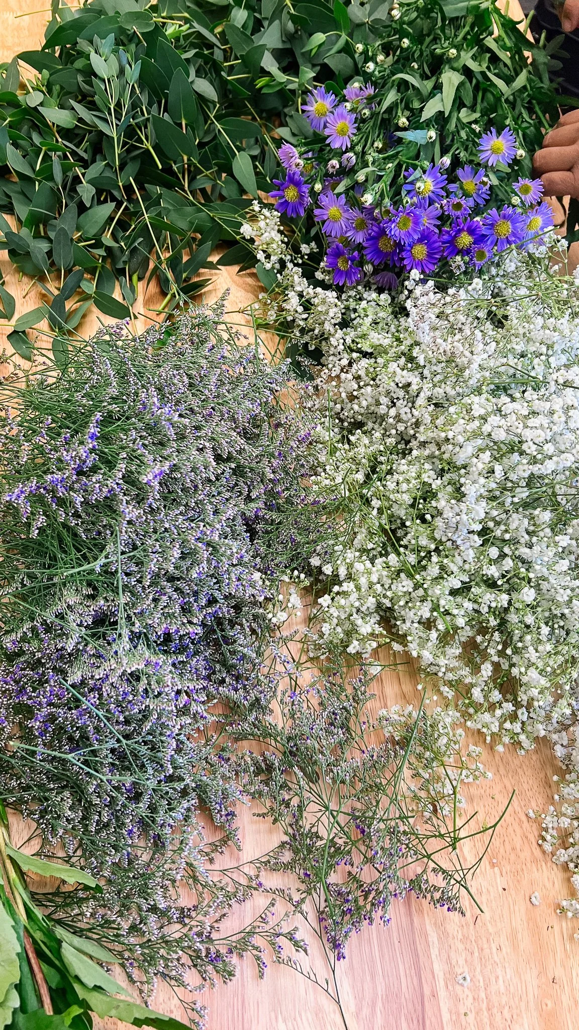 Bunches of limonium, gypsophila (baby's breath), purple asters, and eucalyptus placed on a wood table surface.