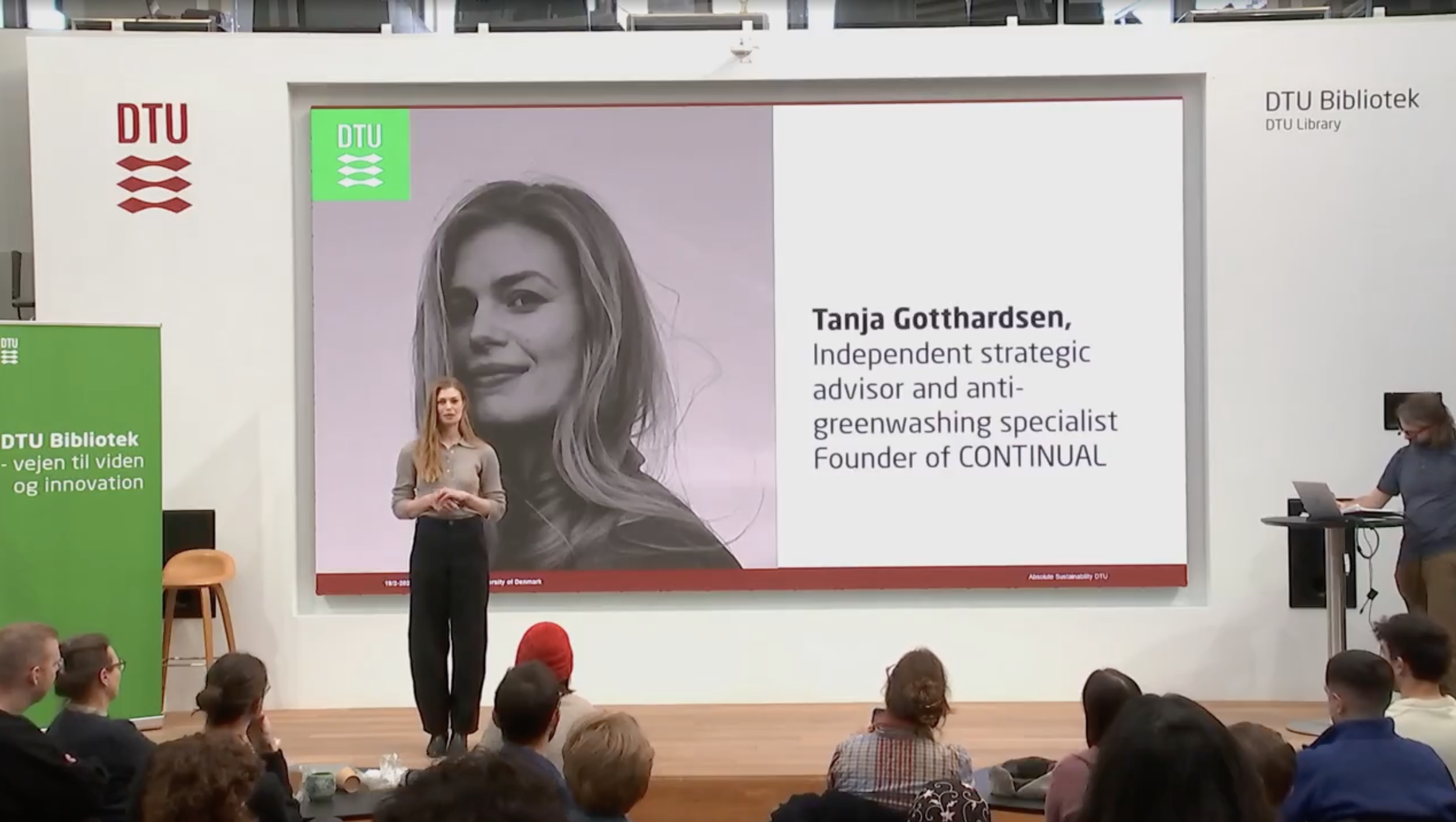 Fremviser, Tanja Gotthardsen, står på en scene foran en stor skærm med hendes navn, titel og foto, med et publikum foran. Der er en grøn DTU-bibliotekskampagne til venstre.