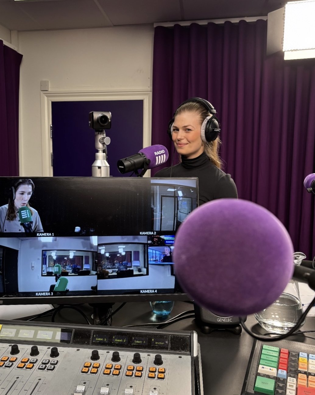 Tanja Gotthardsen i radiostudie med hovedtelefoner foran mikrofon, computer med flere kameraer og redigeringsudstyr. Baggrunden har lilla gardiner.