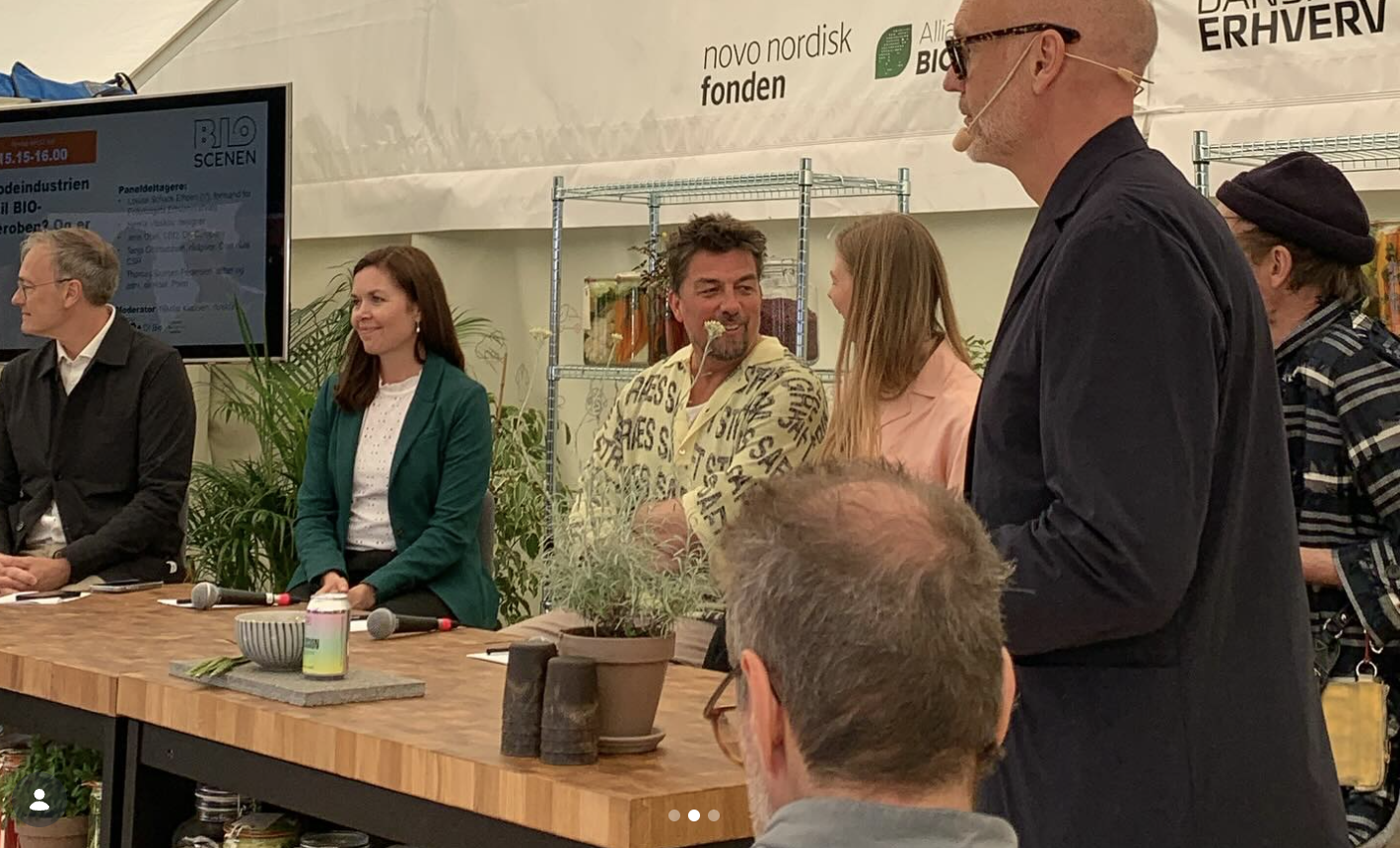 Mænd og kvinder ved et bord til en paneldiskussion, nogle taler, andre lytter, med mikrofoner på bordet, baggrund med plantet og banners.
