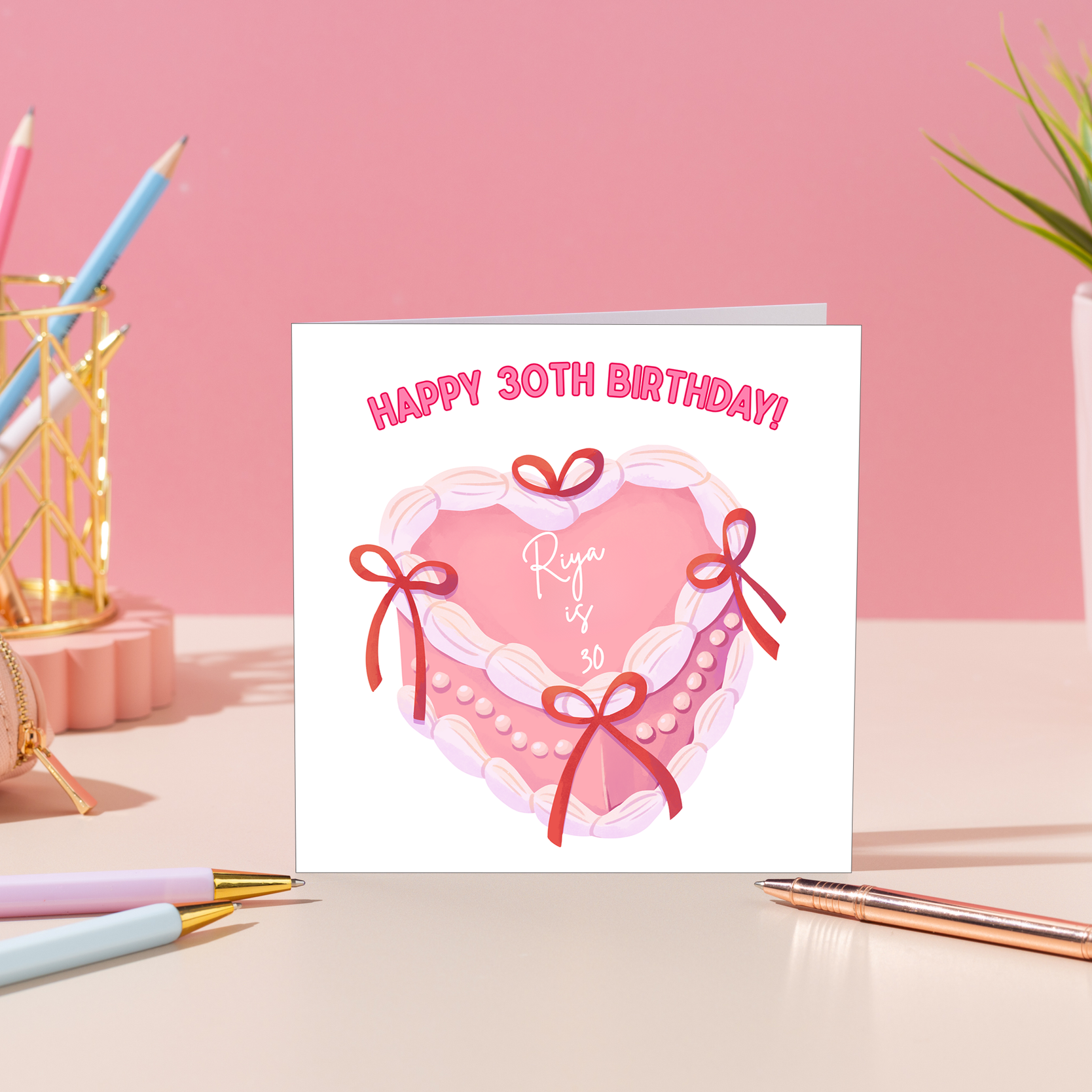 Personalised pink birthday card with heart birthday cake, bows and personalised name and age displayed on a pastel desk
