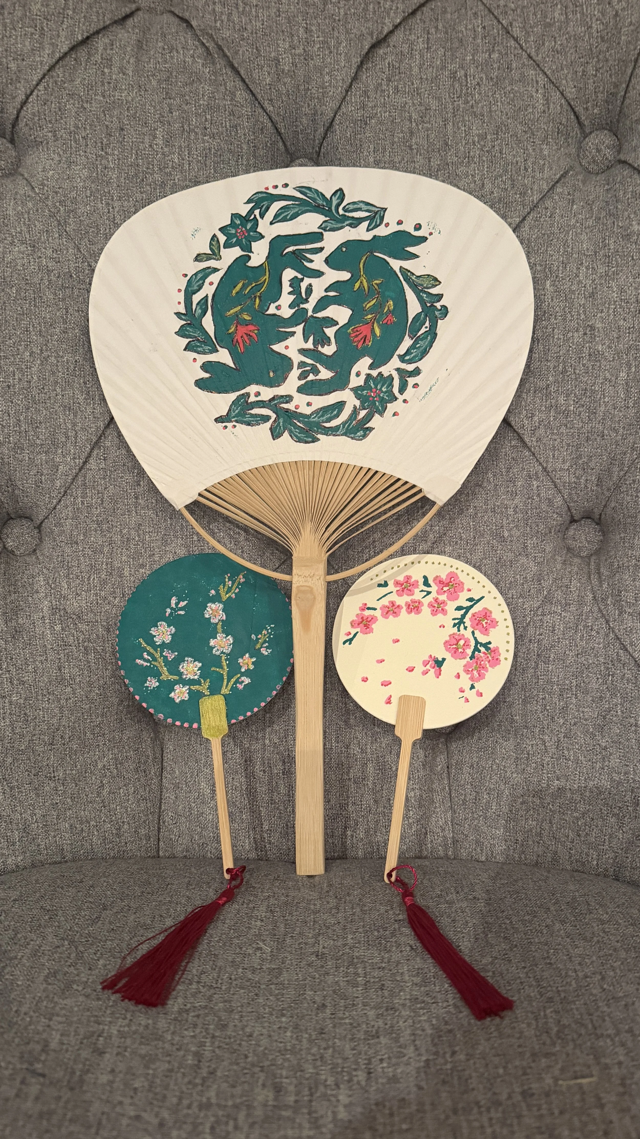 Three decorative handheld fans with floral designs resting against a gray upholstered surface.