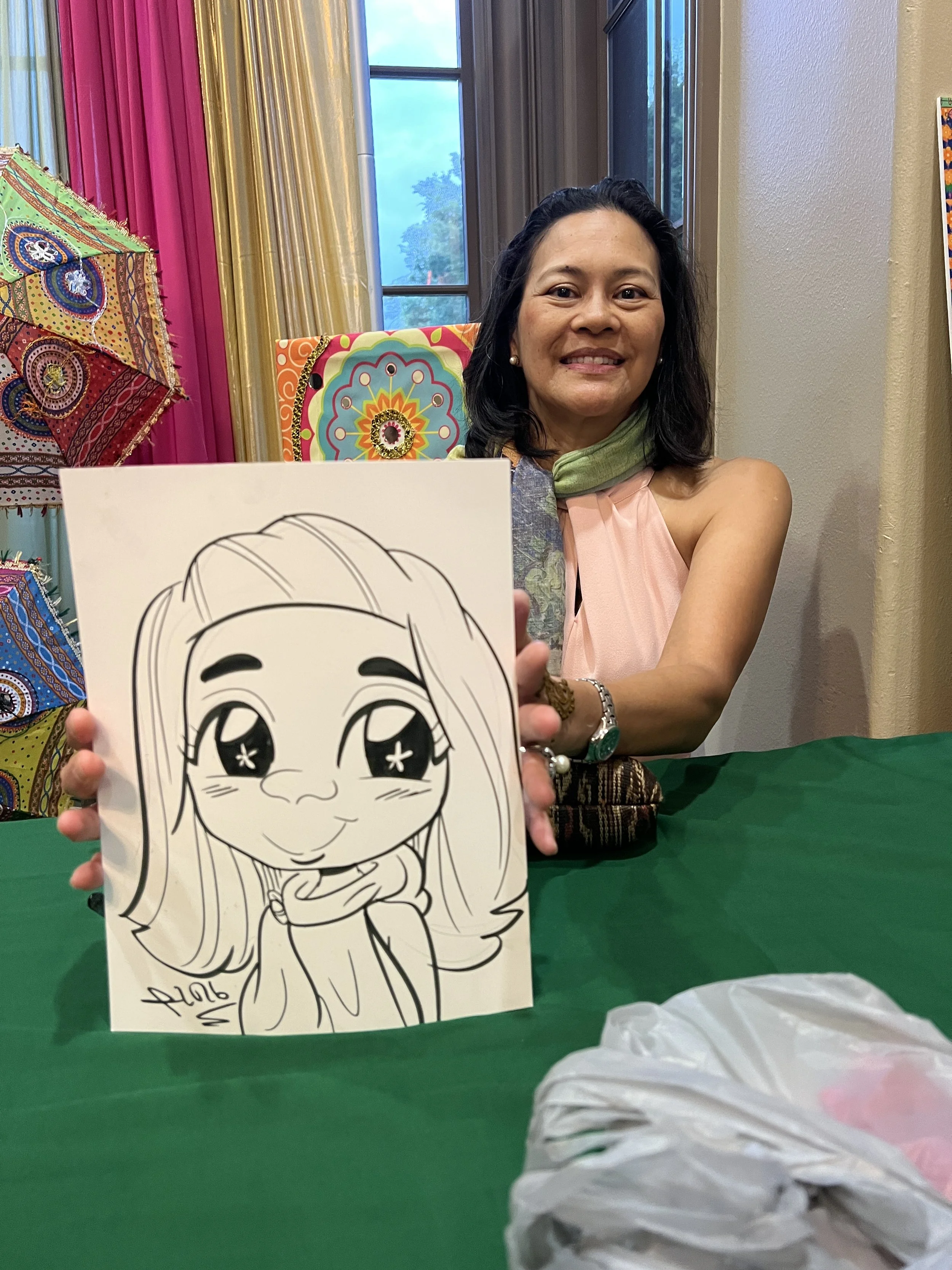 Woman sitting at a table with colorful curtains and window in the background. She is holding a drawing of a cartoon girl with long hair, big eyes, and a cheerful expression, signed with the name 'Alora' and the year 2016.