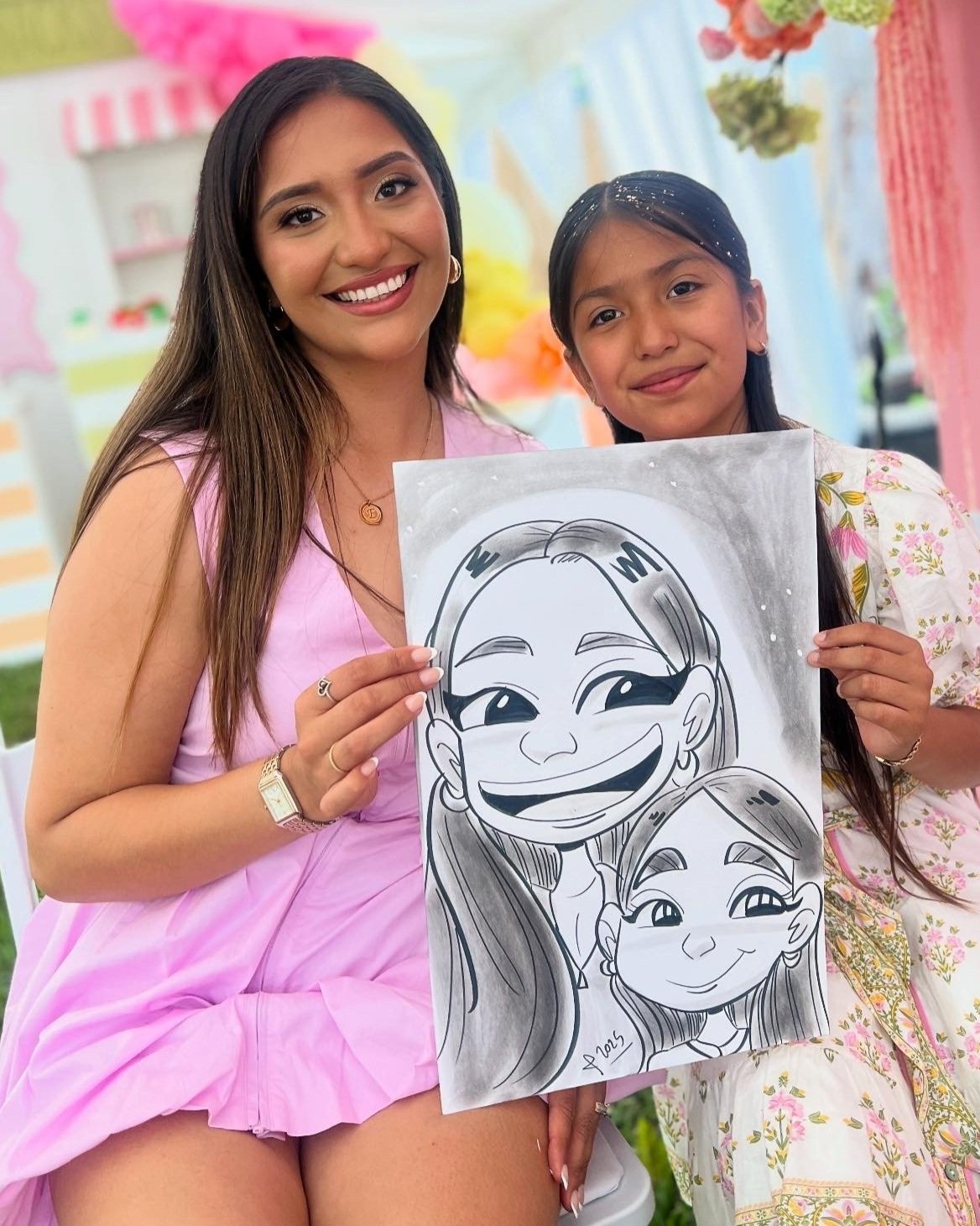 Two women smiling and holding a cartoon portrait drawing of themselves, with a decorated outdoor background.