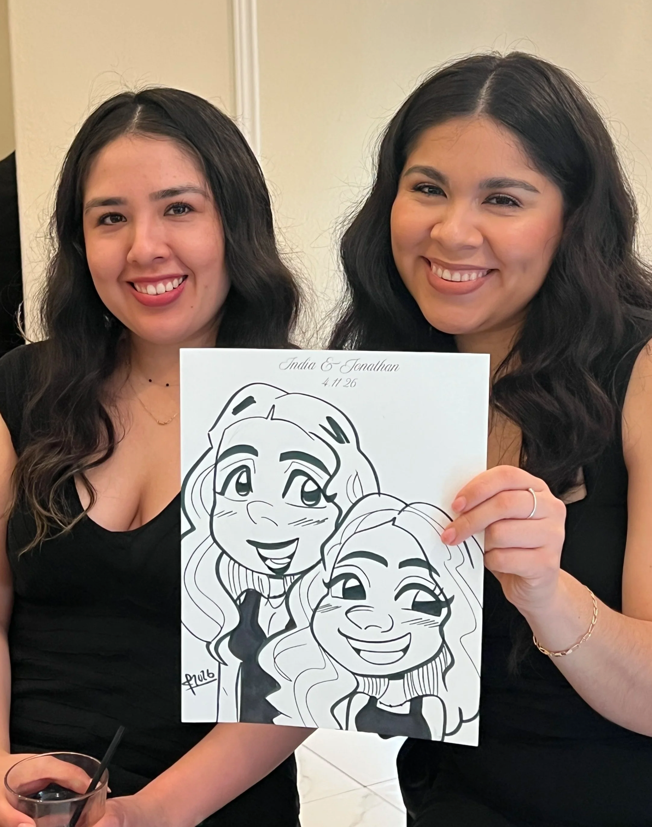 Two women with long dark hair smiling and holding a caricature drawing of themselves, with a white background and part of a table visible in the foreground.