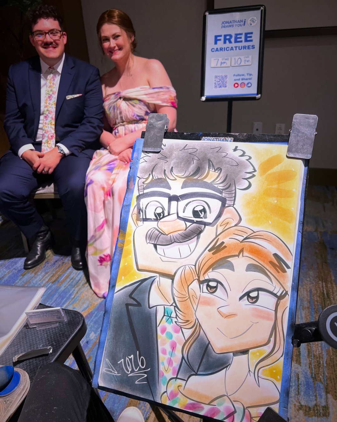 A caricature drawing of a man with glasses and a mustache and a woman with wavy hair, winking, on an easel in front of a smiling man and woman sitting side by side at a table.