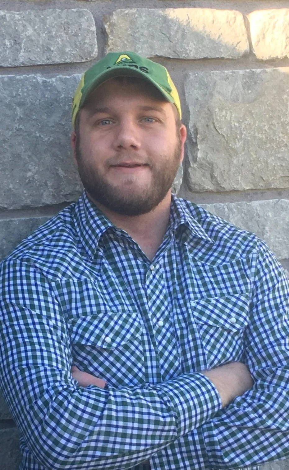 A man with a beard wearing a green and yellow farm cap and a blue plaid shirt, standing with arms crossed in front of a stone wall.