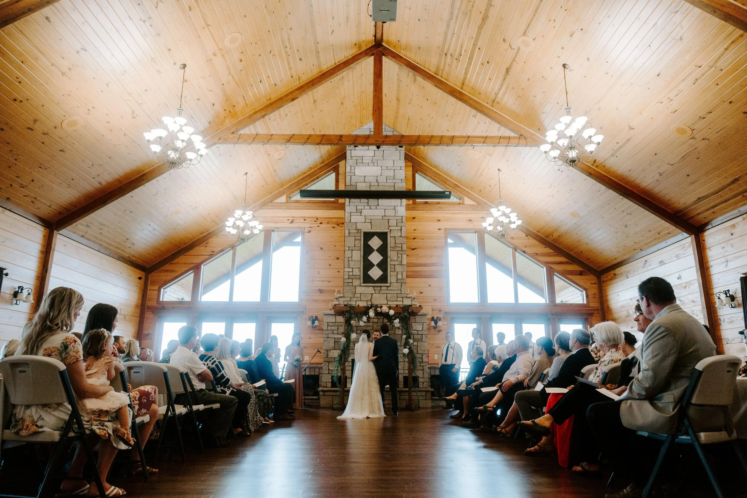 A wedding ceremony taking place in a modern wooden A frame banquet hall with guests seated on either side, the bride and groom standing before a large stone fireplace, with bright large windows behind them.
