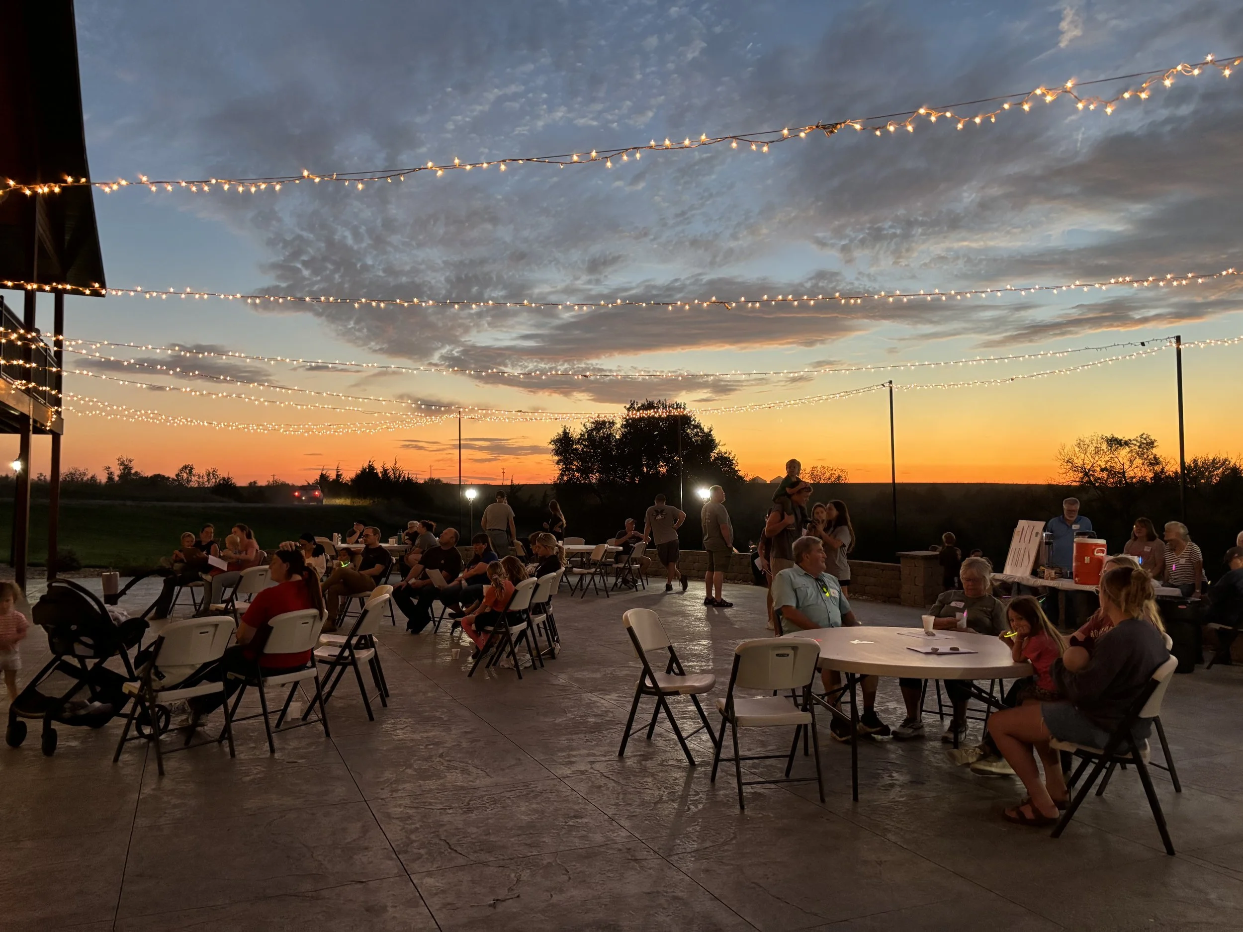Guests at tables on patio with string lights above and orange sunset at Clay Center, KS venue.