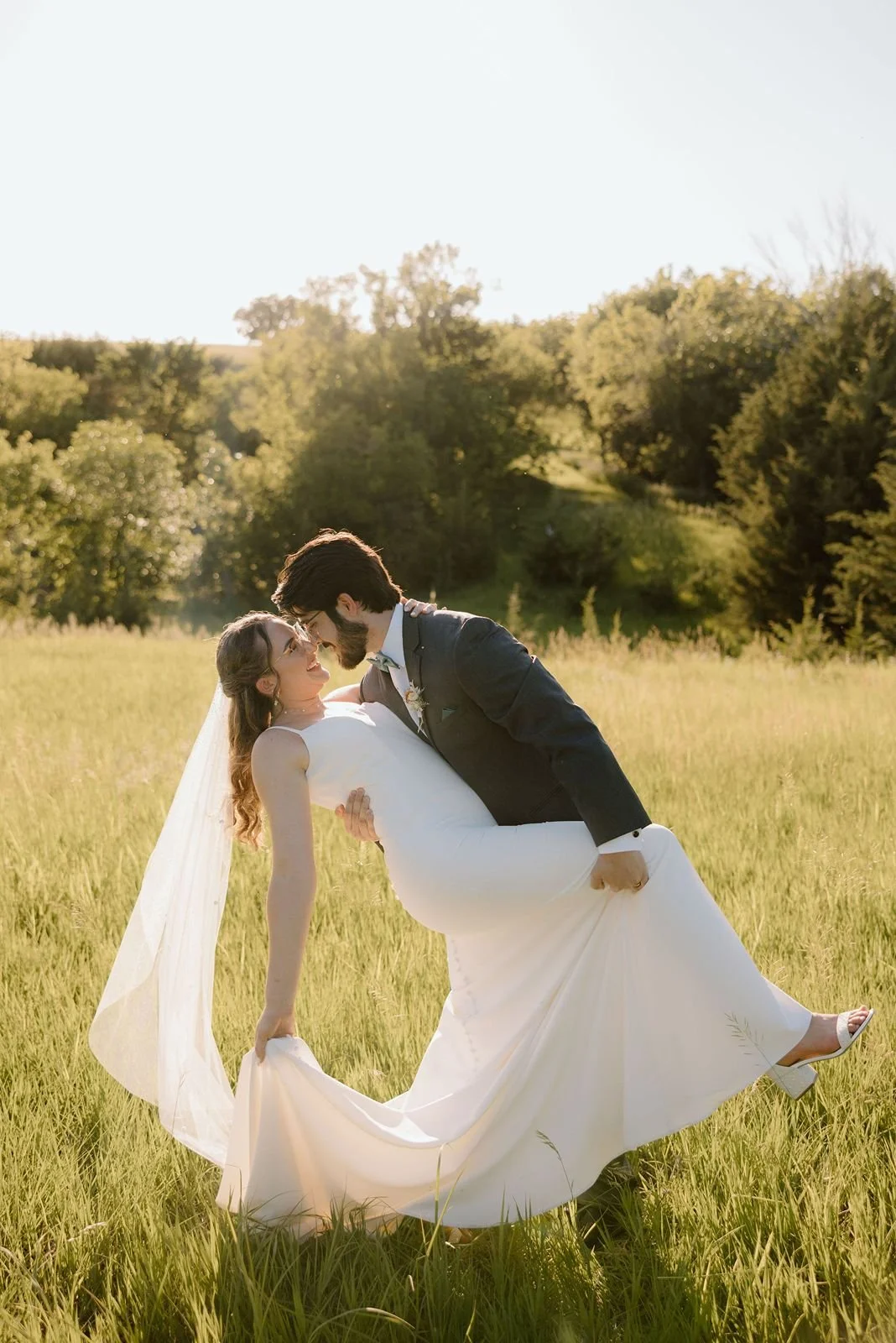 A newlywed couple sharing a dance in a grassy field during sunset, with trees and hills in the background.
