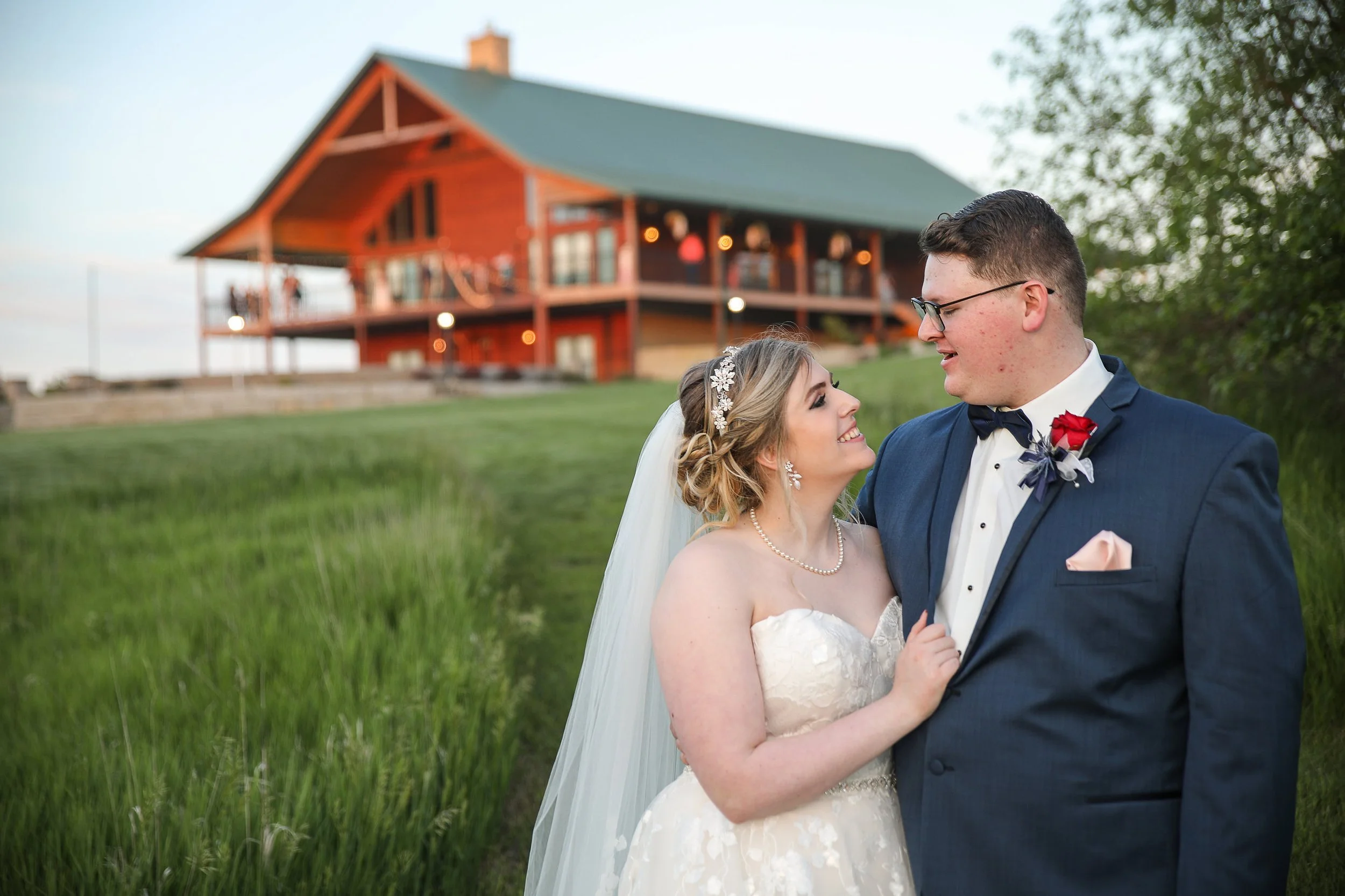 Bride and groom smiling at each other outdoors with a large wooden mountain-type venue in the background.