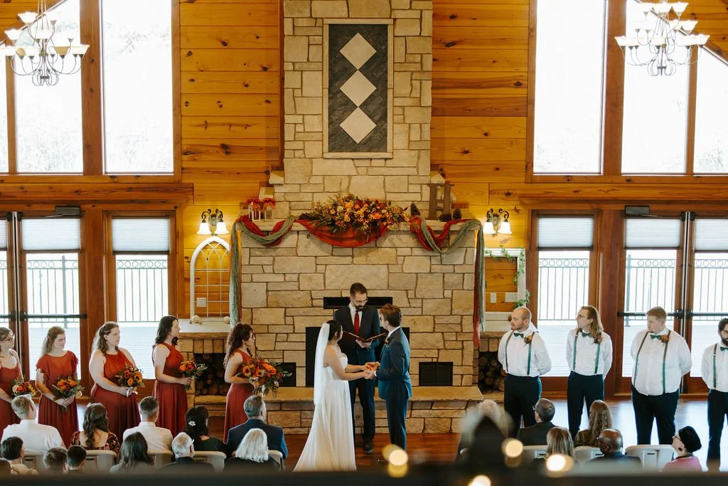 Couple exchanging vows with tall limestone fireplace and tall windows and bridal party.