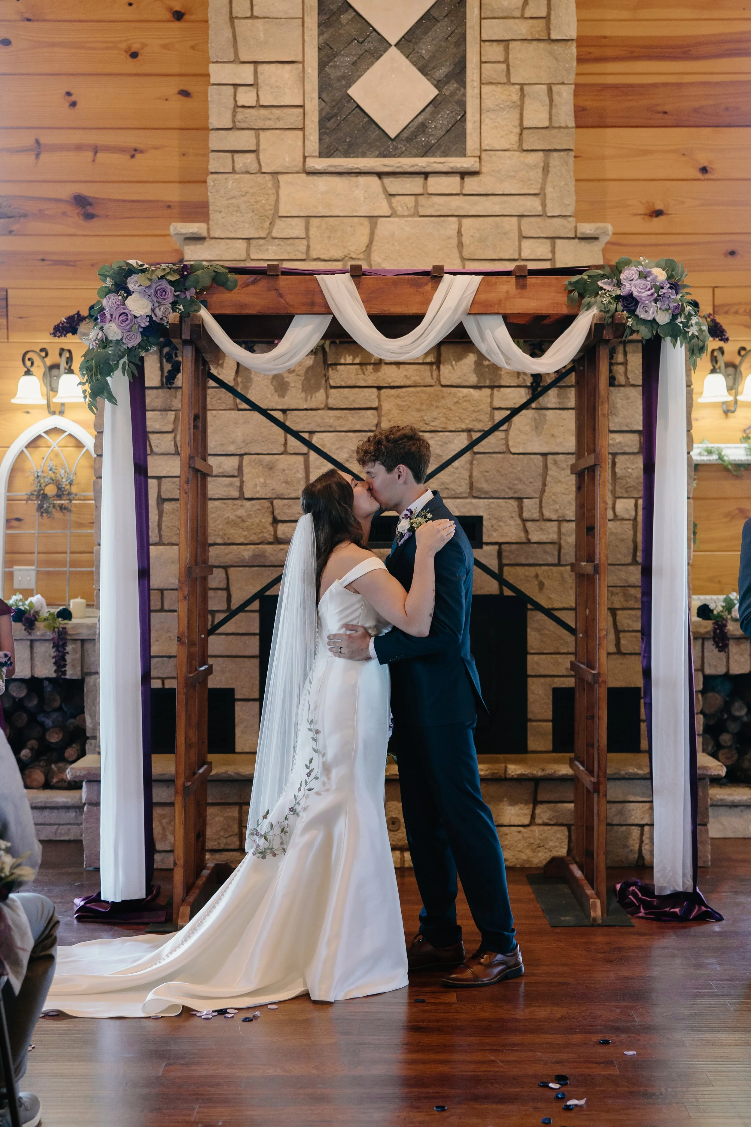 wedding couple kissing in front of wood decorated arbor in front of fireplace inside wood lodge