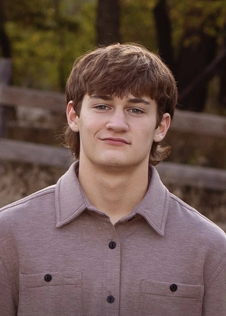 Young man with brown hair outdoors, wearing a gray button-up shirt, with a wooden fence and trees in the background.