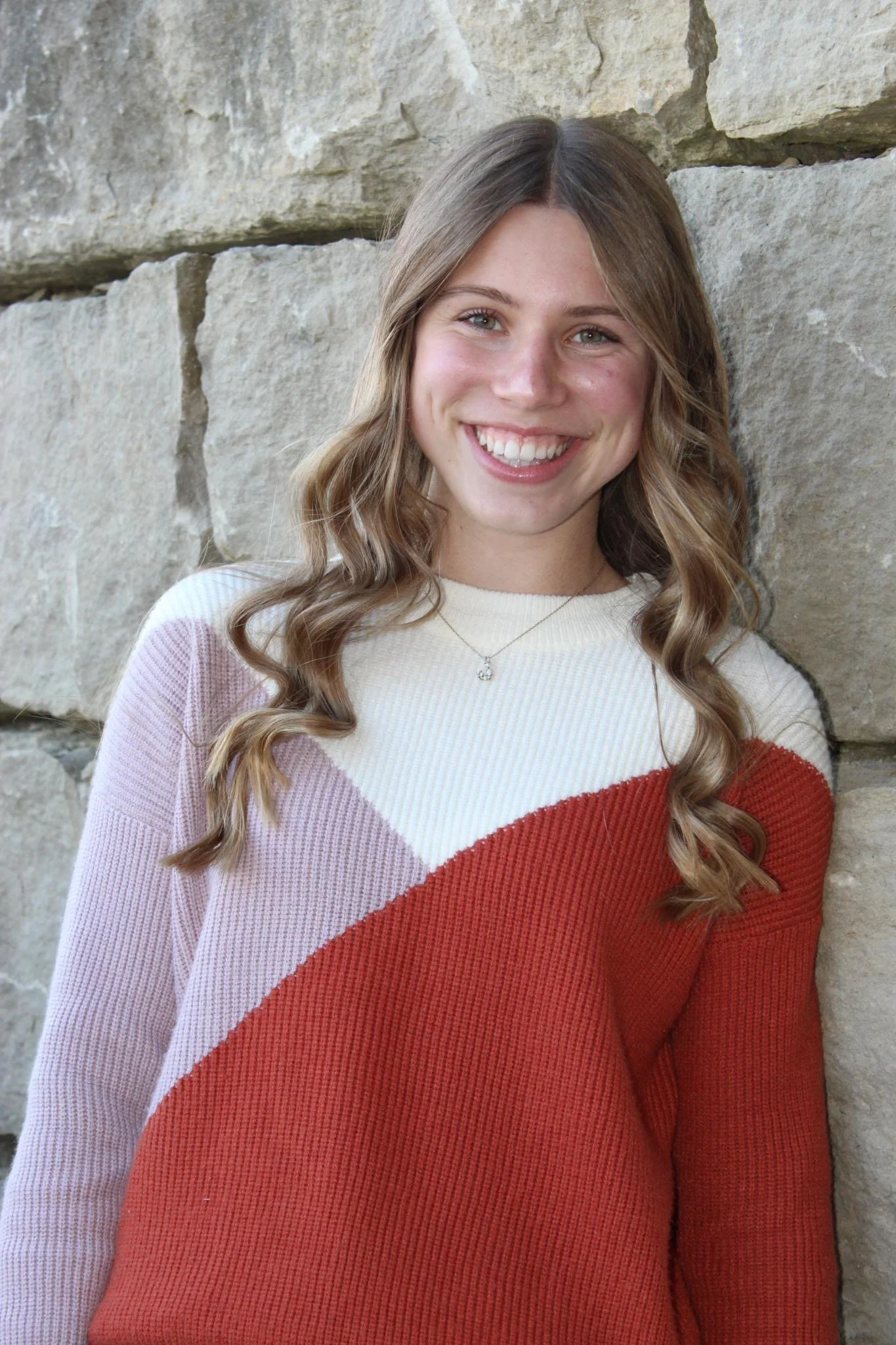 A young woman with long, wavy brown hair smiling, standing against a stone wall, wearing a color-block sweater with cream, pink, and red sections, and a delicate necklace.