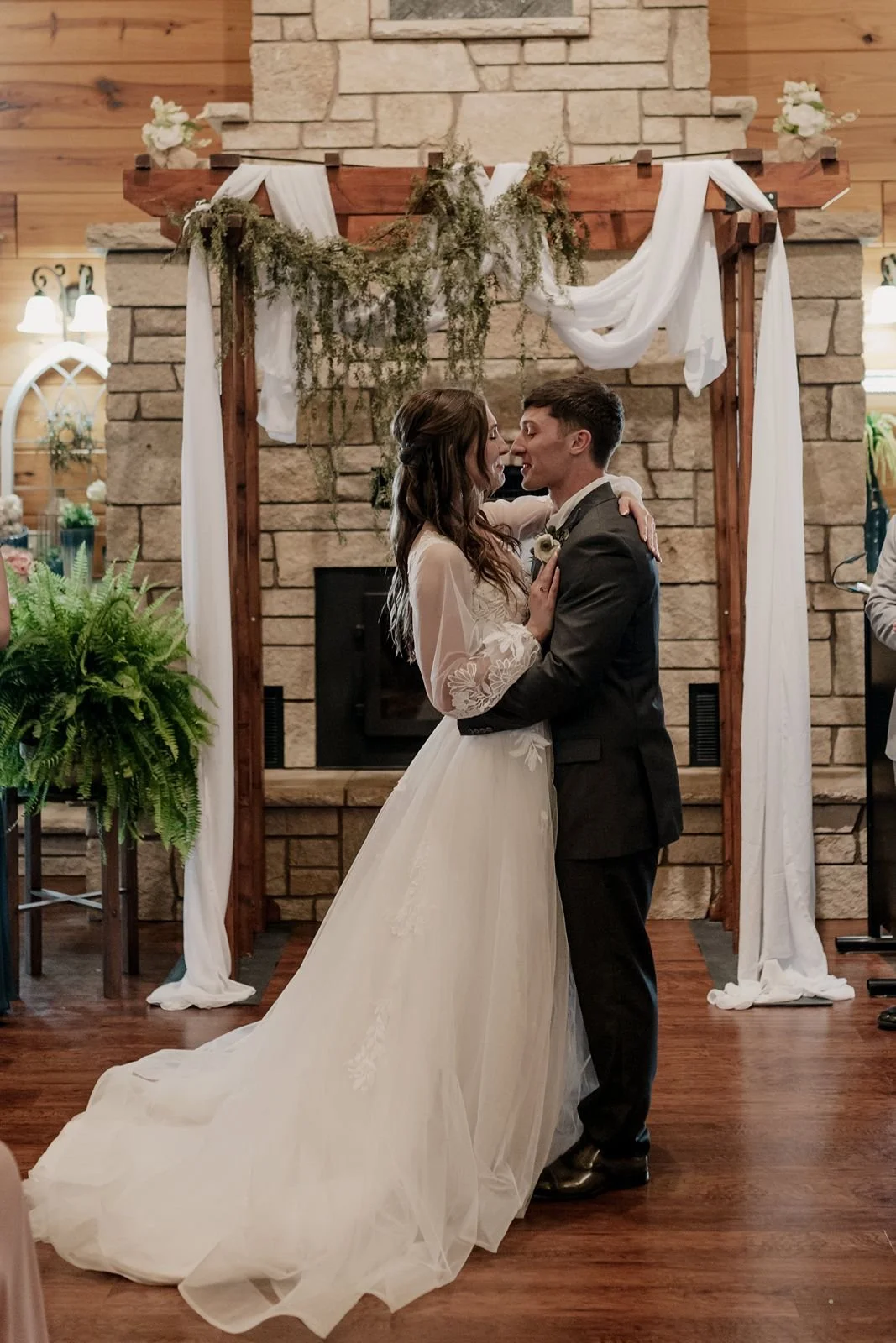 A bride and groom share a dance during their wedding at an indoor venue with a stone fireplace and wooden accents. The bride wears a long white gown with lace sleeves, and the groom wears a dark suit.