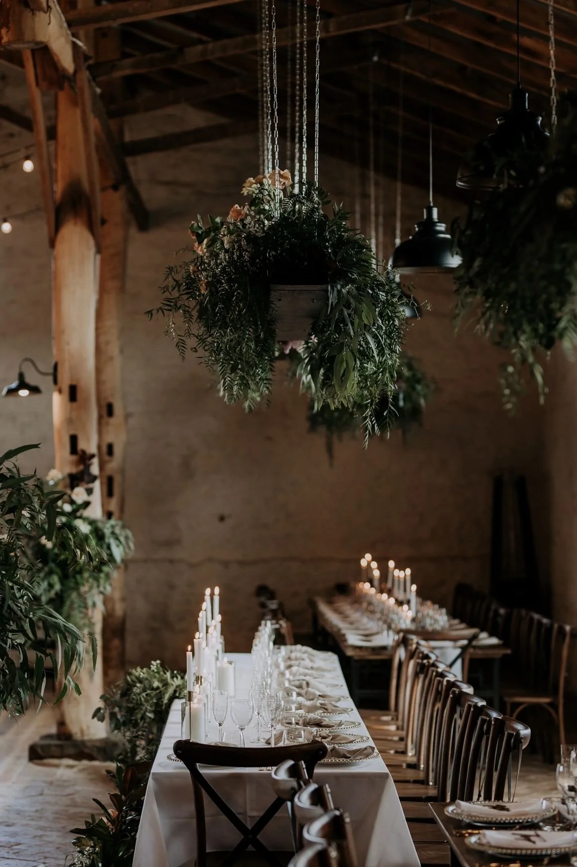 A rustic indoor dining setup with a long table decorated with white tablecloth, candles, and elegant glassware, surrounded by wooden chairs. Above, hanging greenery and flowers in pots add to the natural ambiance, with wooden beams and industrial-sty
