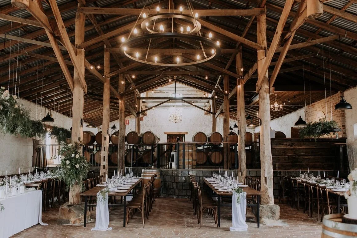 Indoor wedding reception with long dining tables, elegant place settings, and floral centerpieces, in a rustic barn with wooden beams and wine barrels in the background.