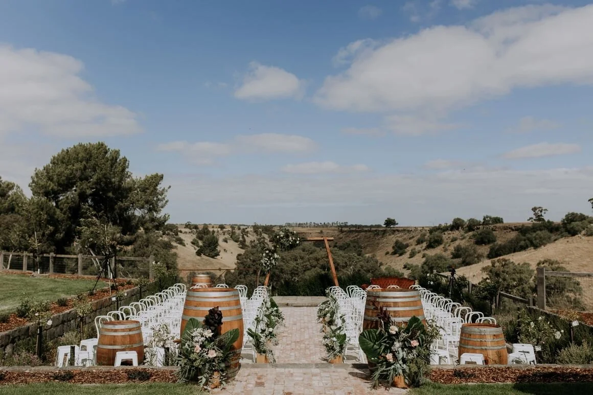 Outdoor wedding ceremony setup with white chairs, decorated barrels, and floral arrangements on a stone-paved aisle, overlooking a scenic landscape of hills and trees under a partly cloudy sky.
