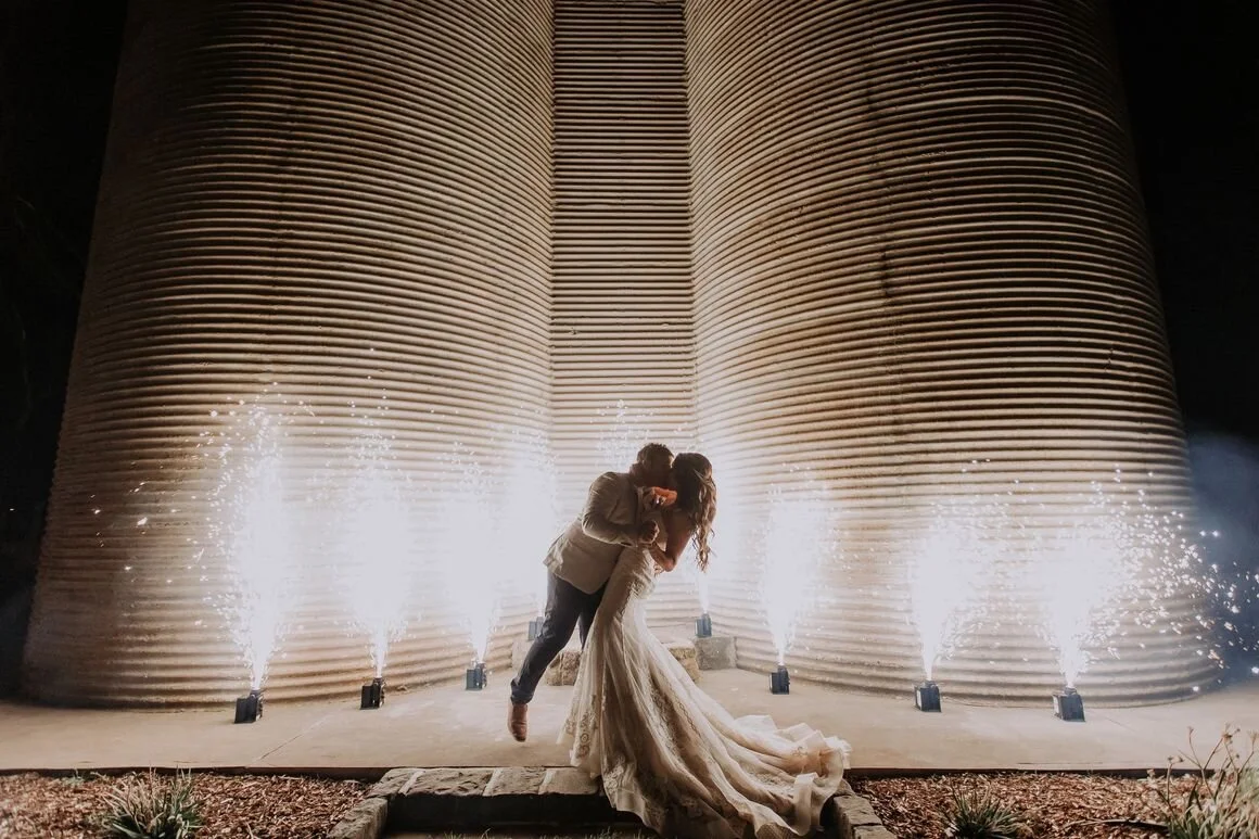 A bride and groom dancing in front of large industrial silos with fireworks at night.