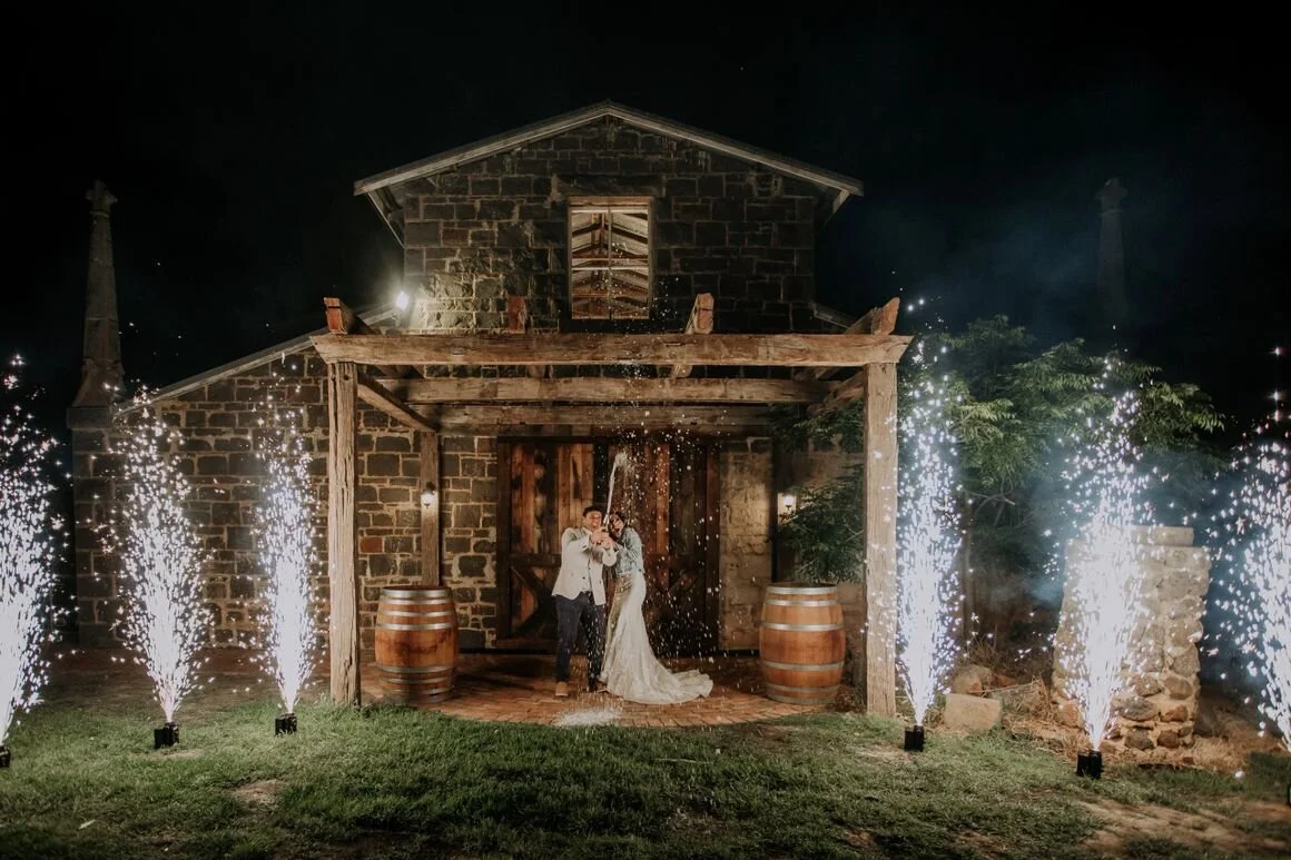 A couple celebrating a wedding at night with sparklers, standing in front of a rustic stone and wood building with barrel decorations, surrounded by fireworks.