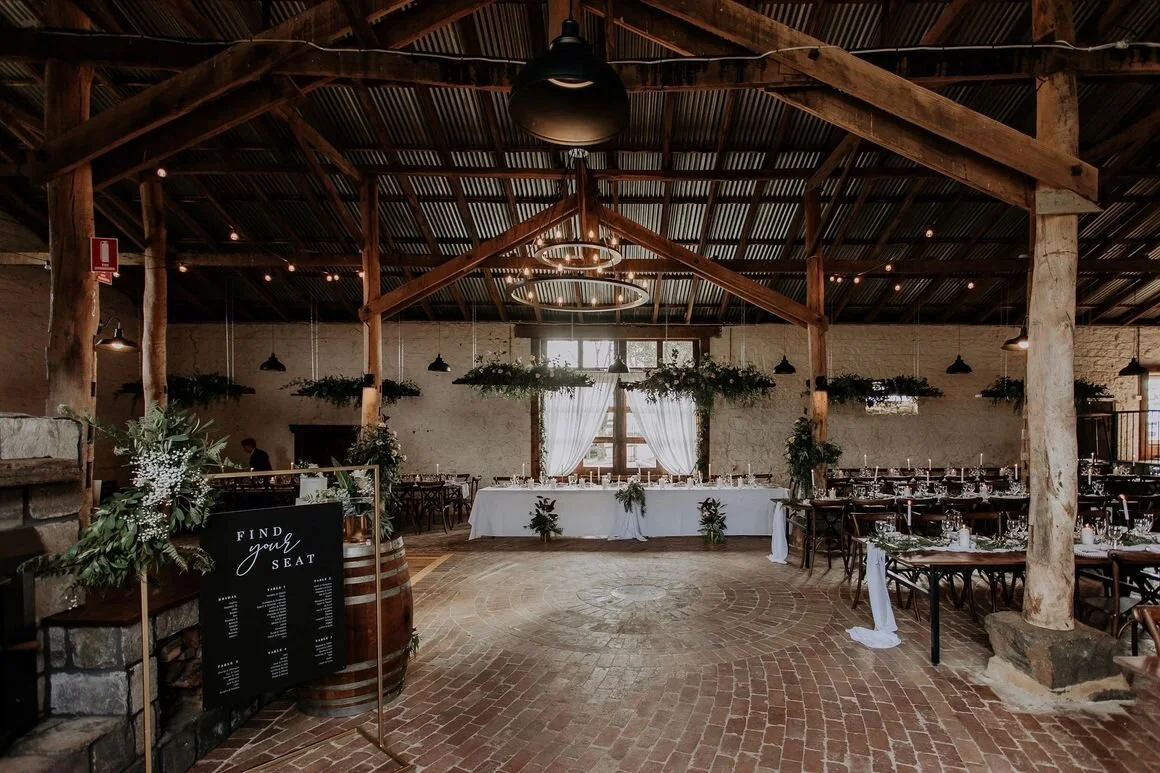 A rustic wedding reception venue with wooden beams, hanging greenery, round tables with white tablecloths, and a long head table at the front. A sign asks guests to find their seat, and the space is decorated with drapes and natural decor.