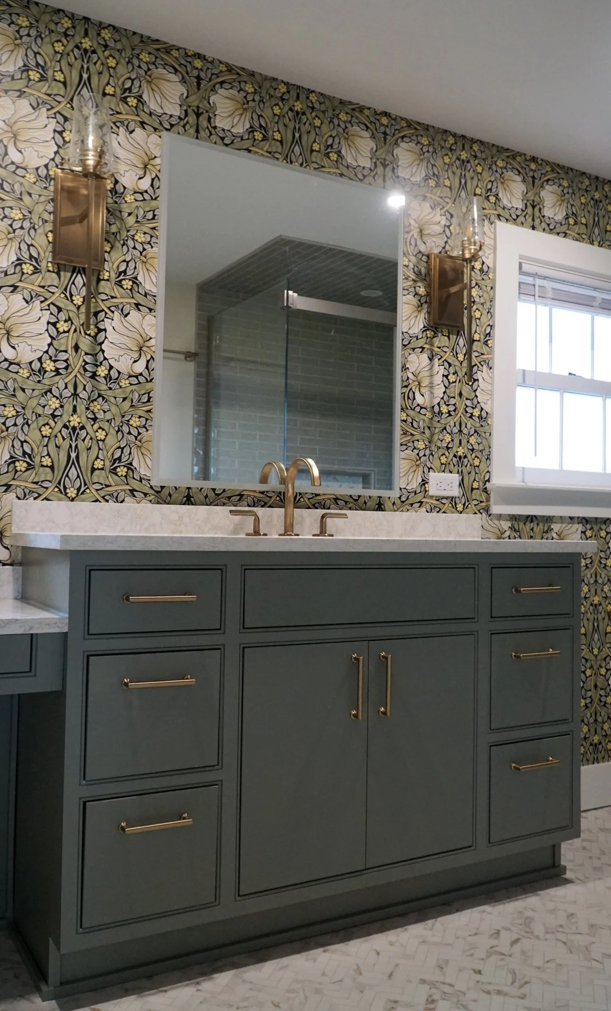 A bathroom countertop with green cabinets and gold handles. Two light fixtures surround a mirror on the wall which reflects a modern shower in the background. Dramatic green wallpaper with white flowers and black accents.