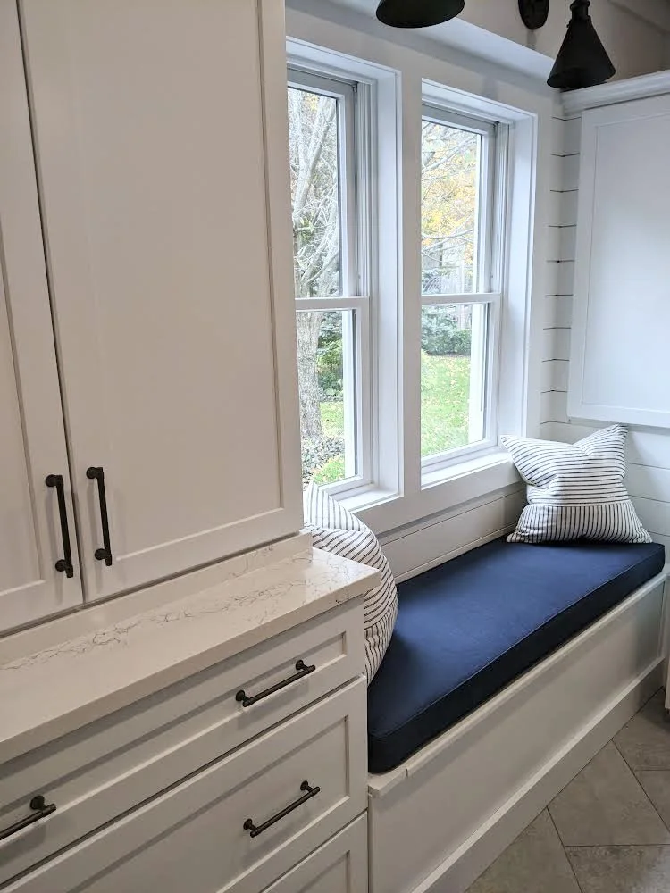 A kitchen nook with a white built-in window seat and navy cushion, striped pillows, and a view of trees outside through two large windows.