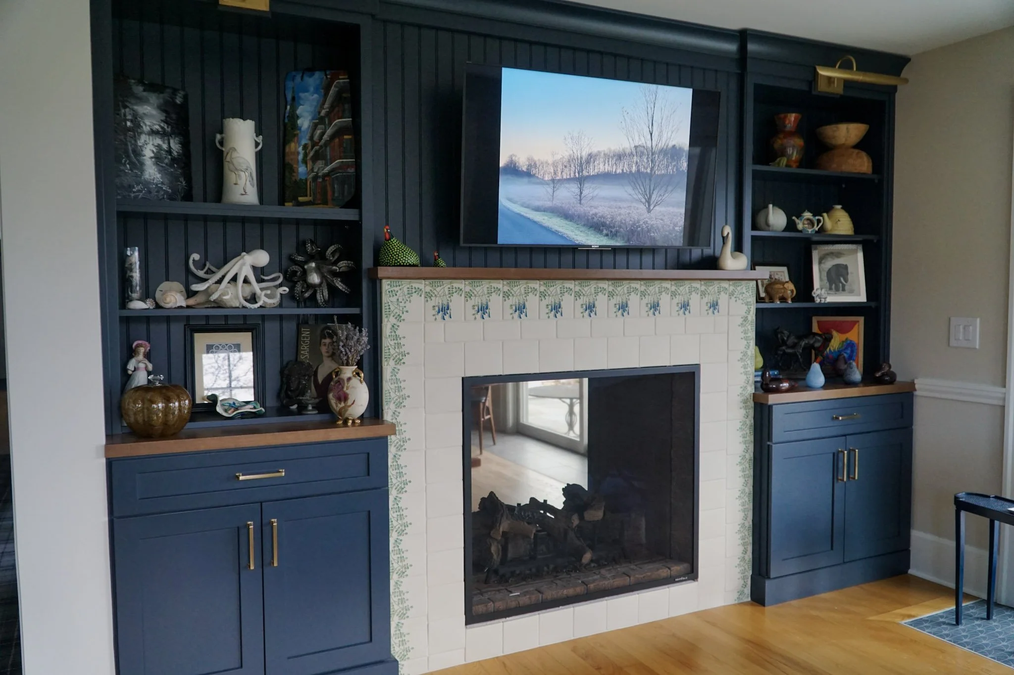 A fire place surrounded by custom painted white tile with blue flowers and green vines. The fireplace looks into the other room and is surrounded by dark blue built in cabinets. A TV sits on top of the fireplace with a nature photo displayed. There a