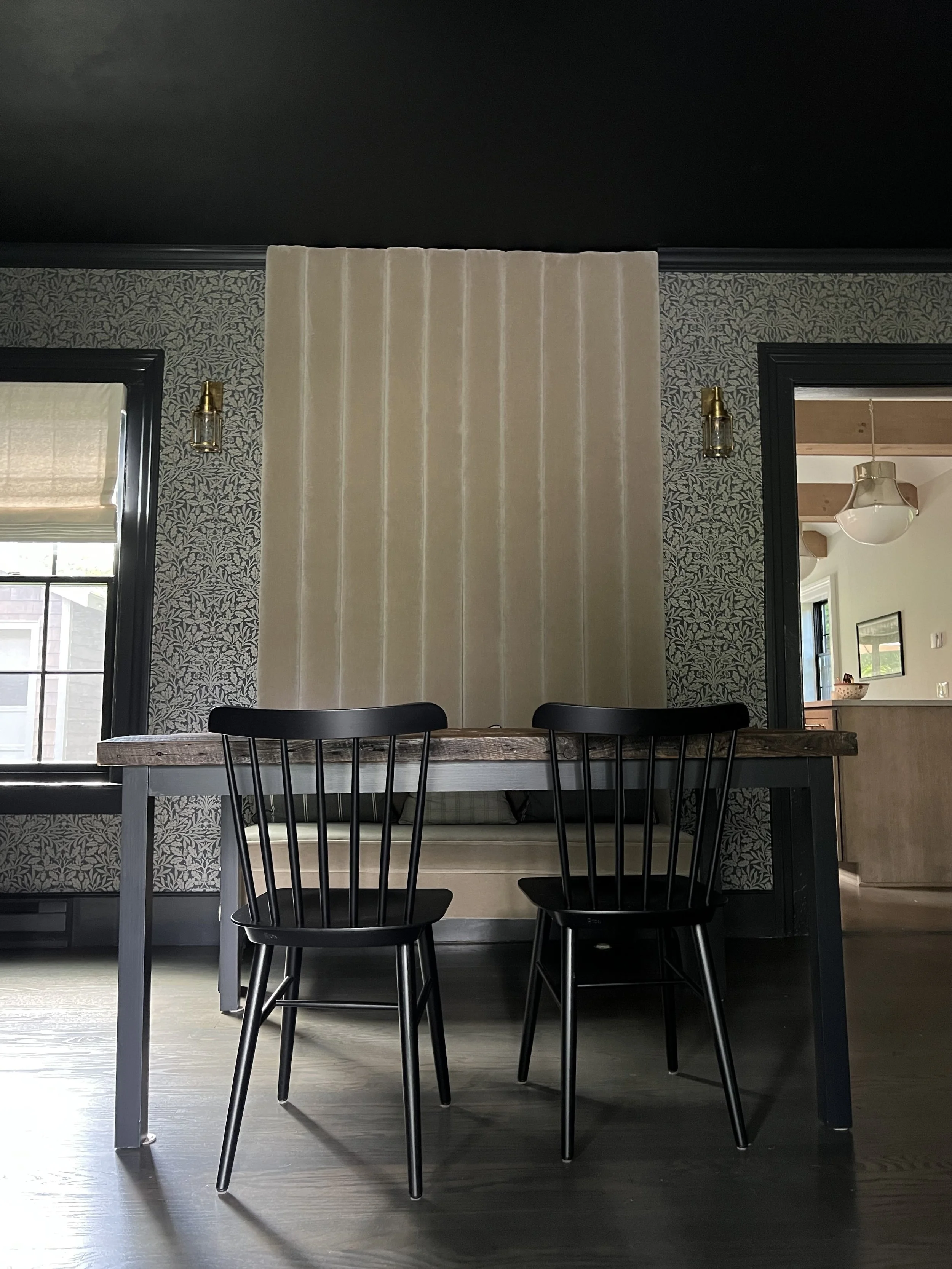Interior dining area with a wooden table, two black chairs, wallpapered walls, a window with blinds, and a decorative wall divider.